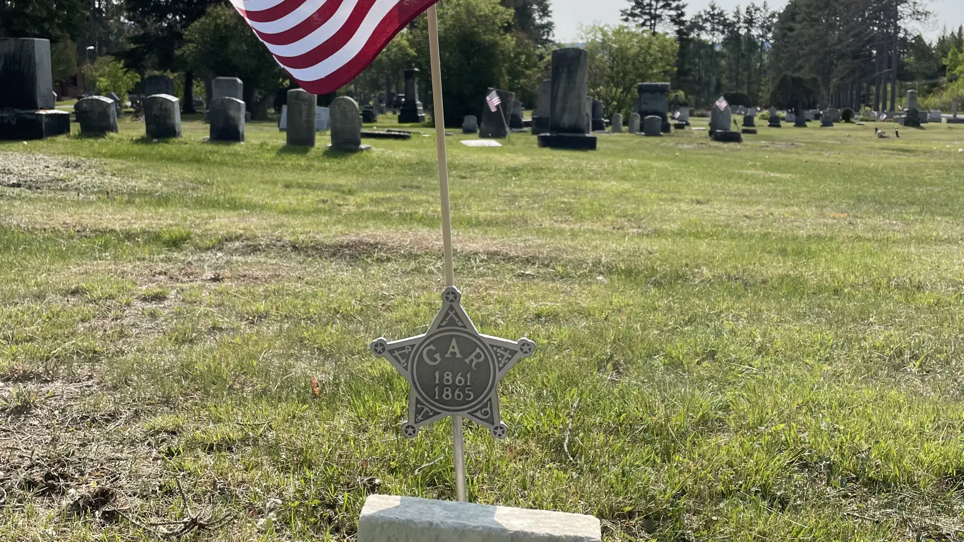 A small gravestone amid green grass with the name William and a metal star stick into the ground behind it that says GAR 1861 and 1865 and above that a small American flag