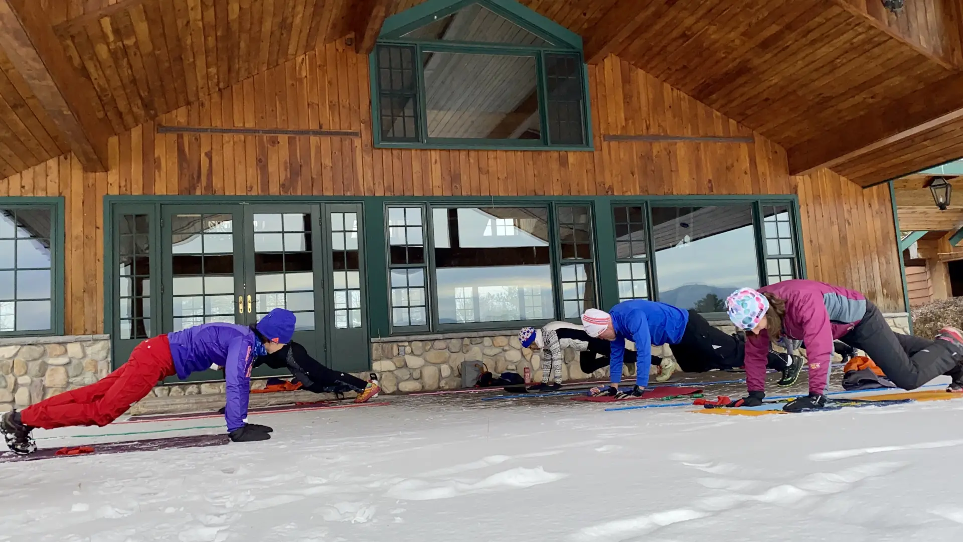 Four people doing planks outside on snow in front of a lodge.