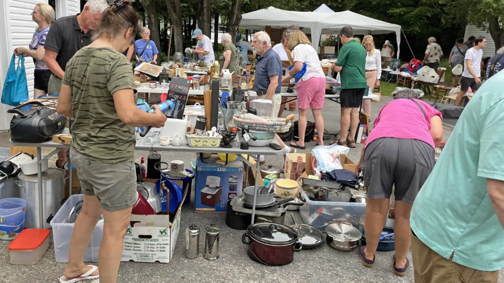 People browse outdoor bazaar sale with items on tables, on the ground, and under pop-up tents
