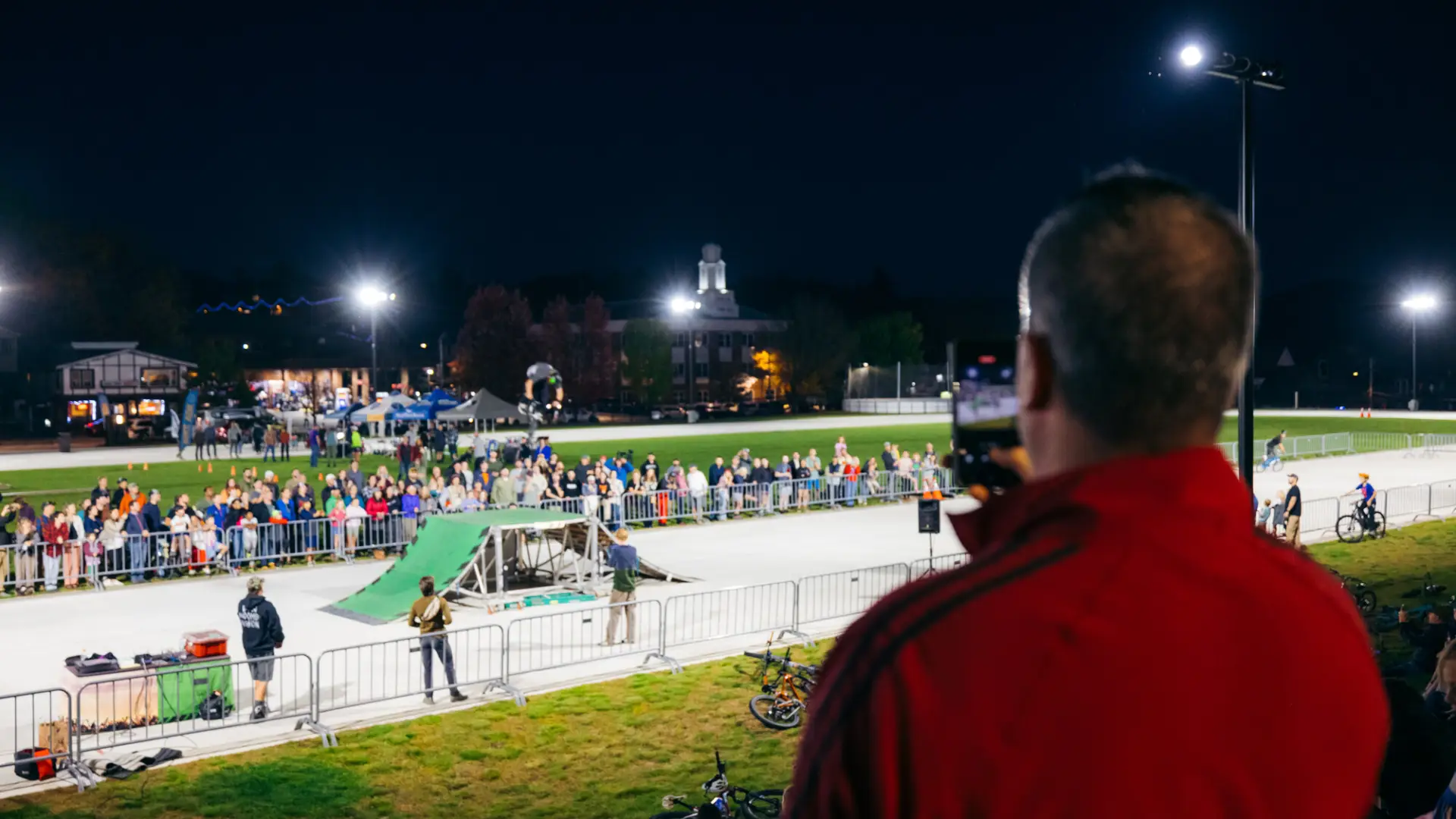 Man takes video of BMX stunt show rider performing a flip off a ramp at the Olympic Oval under the lights at night