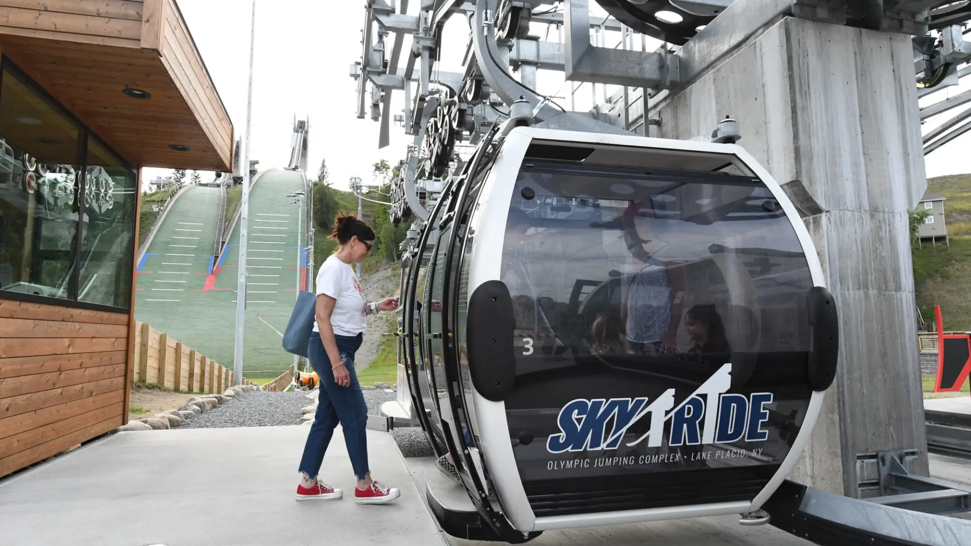 A woman steps into a gondola in front of green ski jumps.