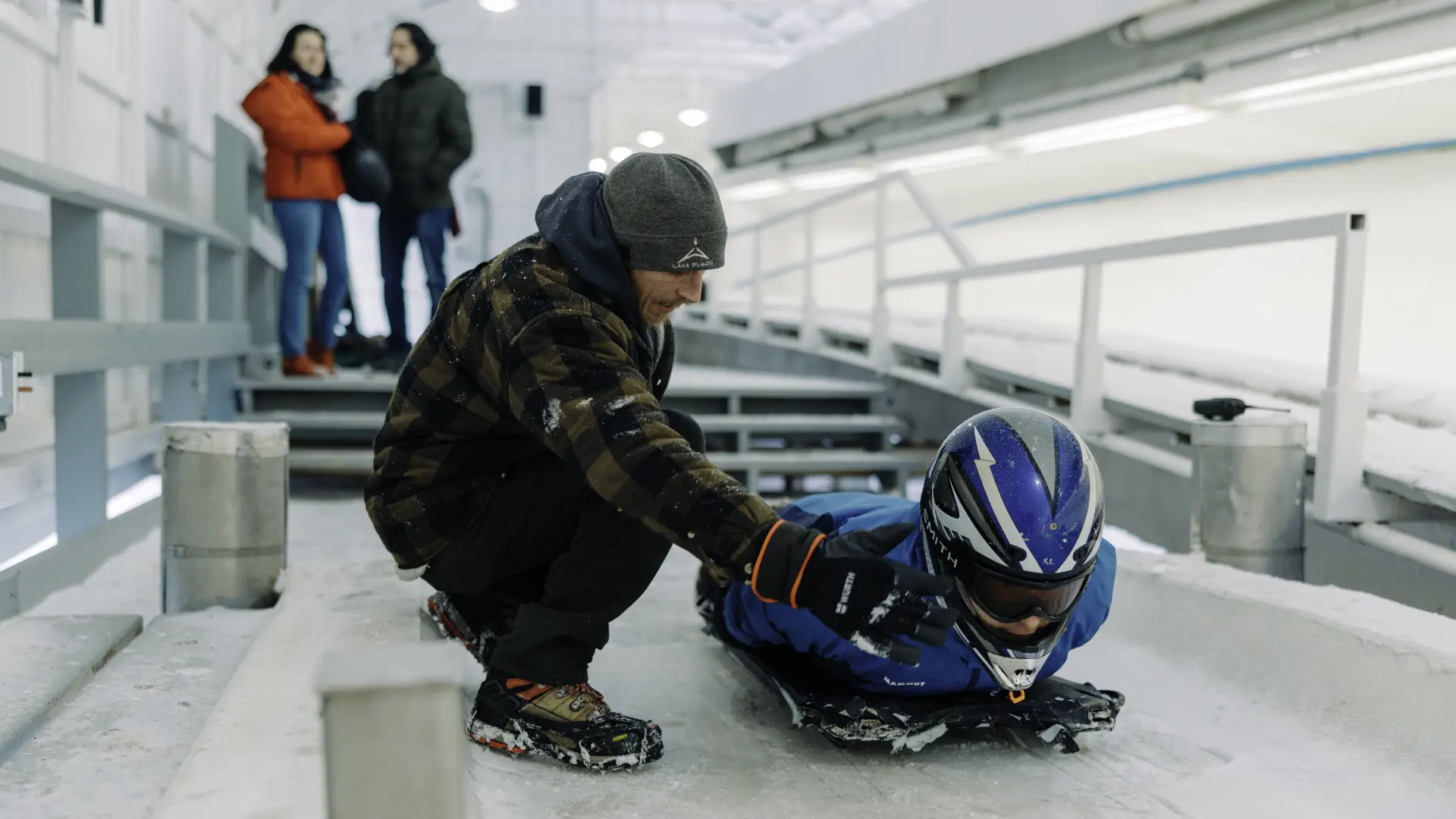 Track worker provides instruction to person in helmet laying on skeleton sled on bobsled, luge, and skeleton track