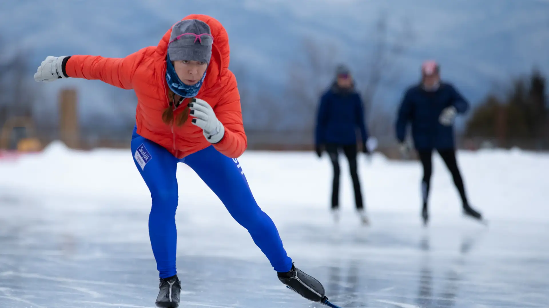 Person in red puffy coat and bright blue spandex pants wearing a hat and white cloves speed skates and a crouched position on an outdoor skating oval. 