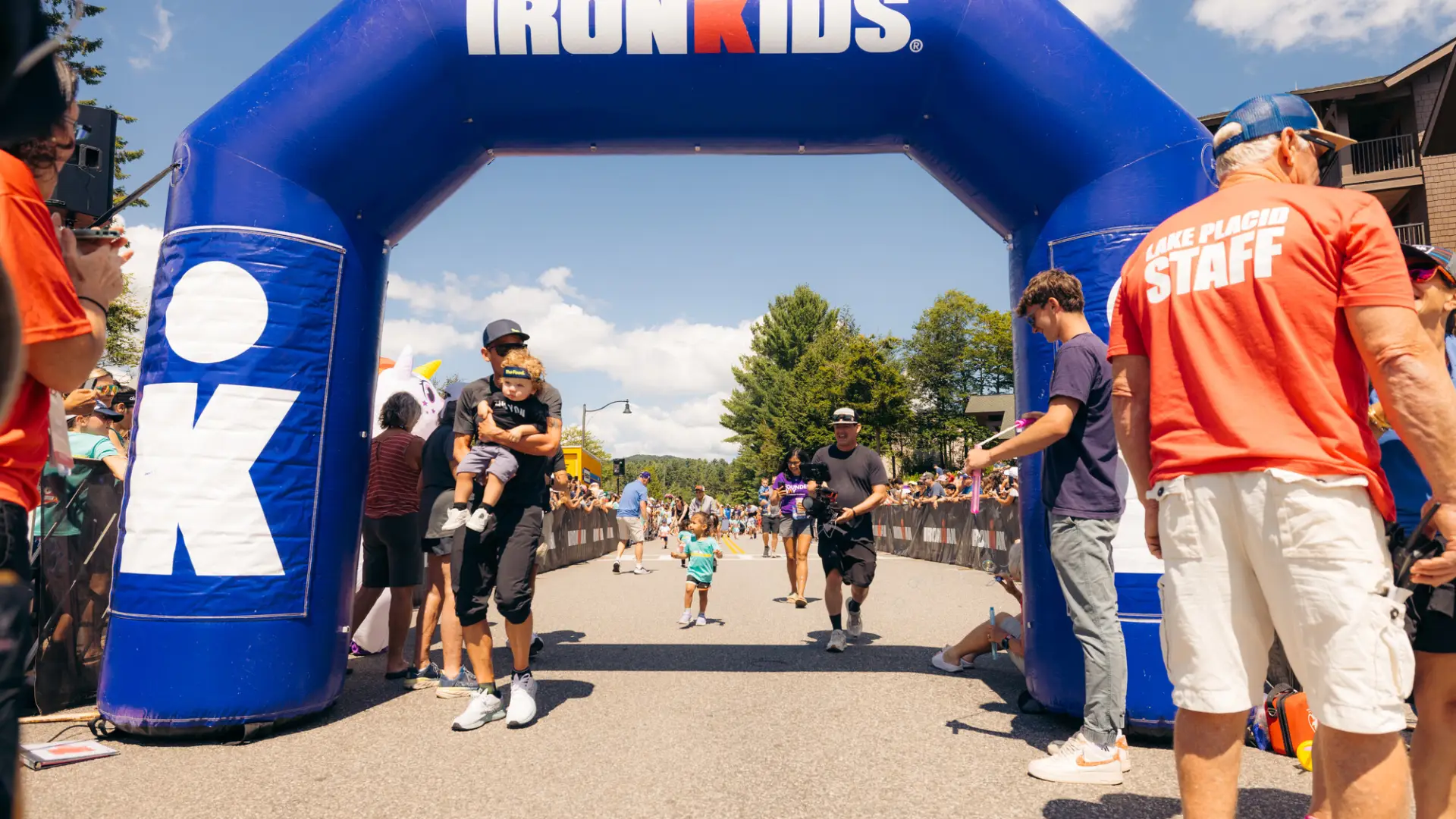 Man holding toddler crosses Ironkids finish line under blue inflatable Ironkids archway