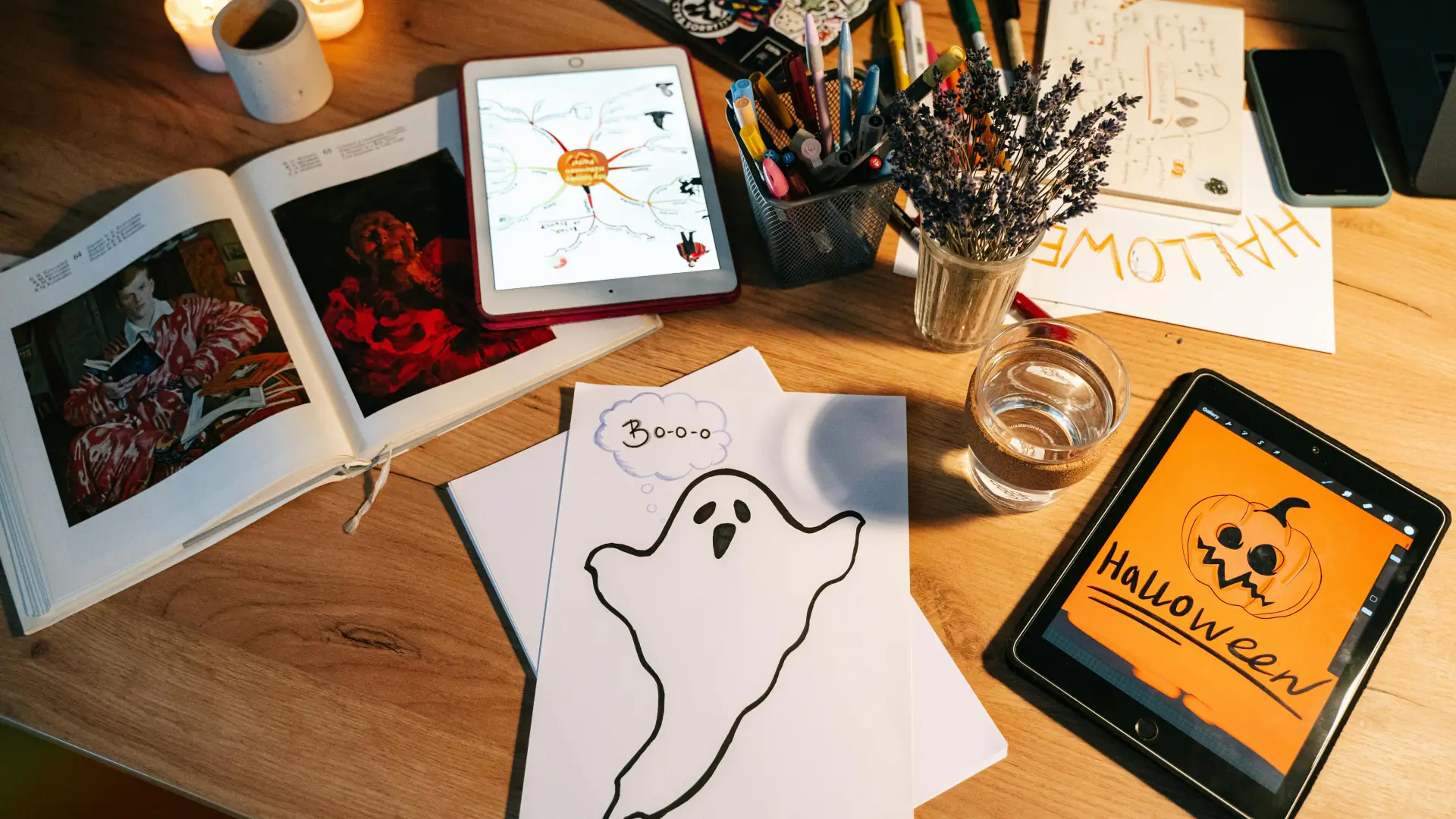 Stock image of table with art supplies, paper with drawing of a ghost, photobooks lying open, and a tablet with a digital halloween drawing