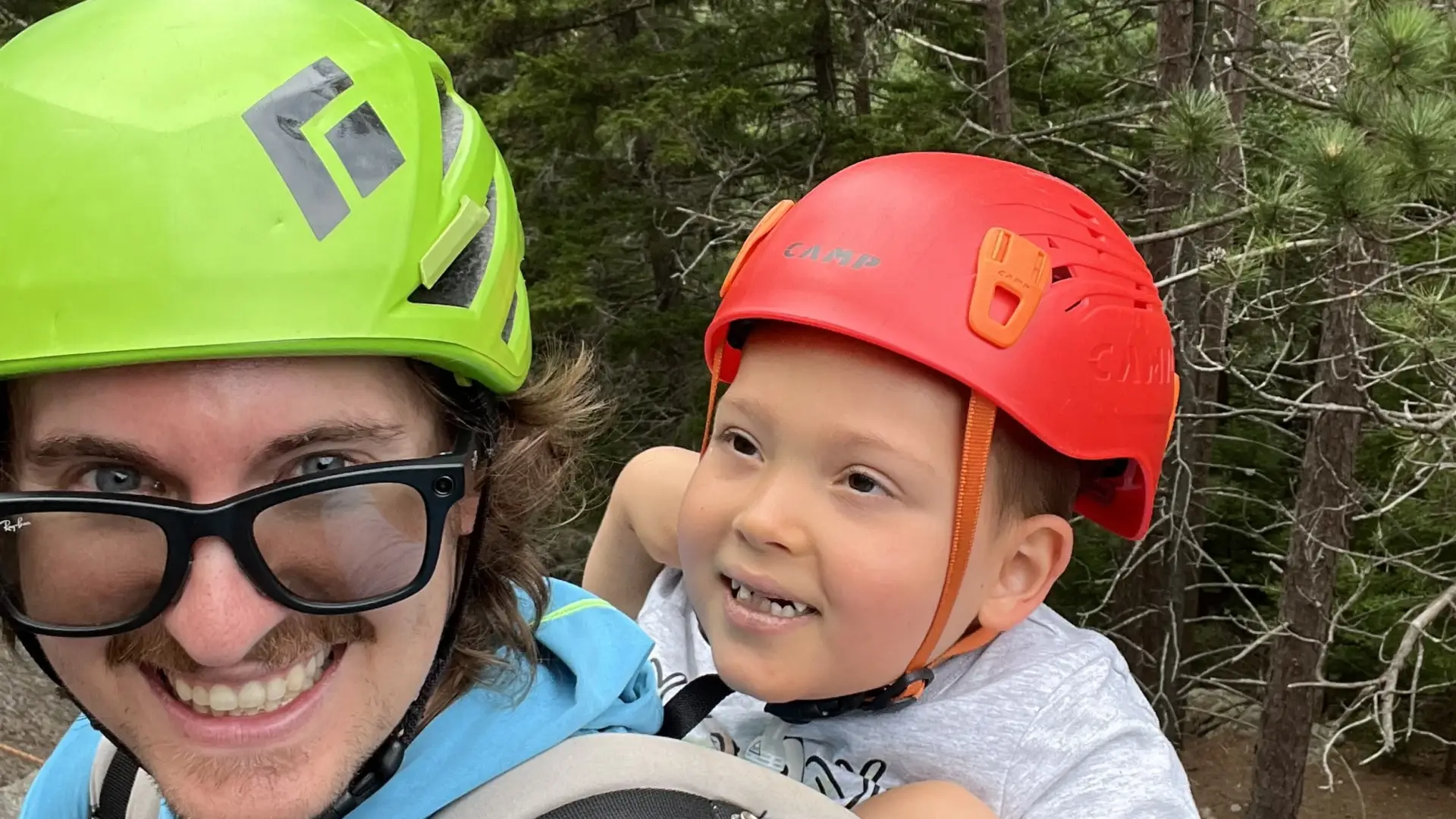 A smiling man faces camera while carrying a young child in a back pack, both donning helmets.