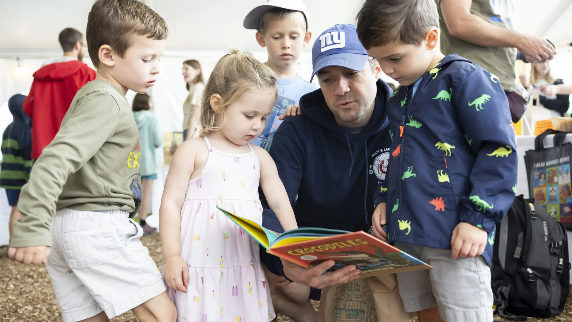 Three small children gather around a man crouching and reading a book