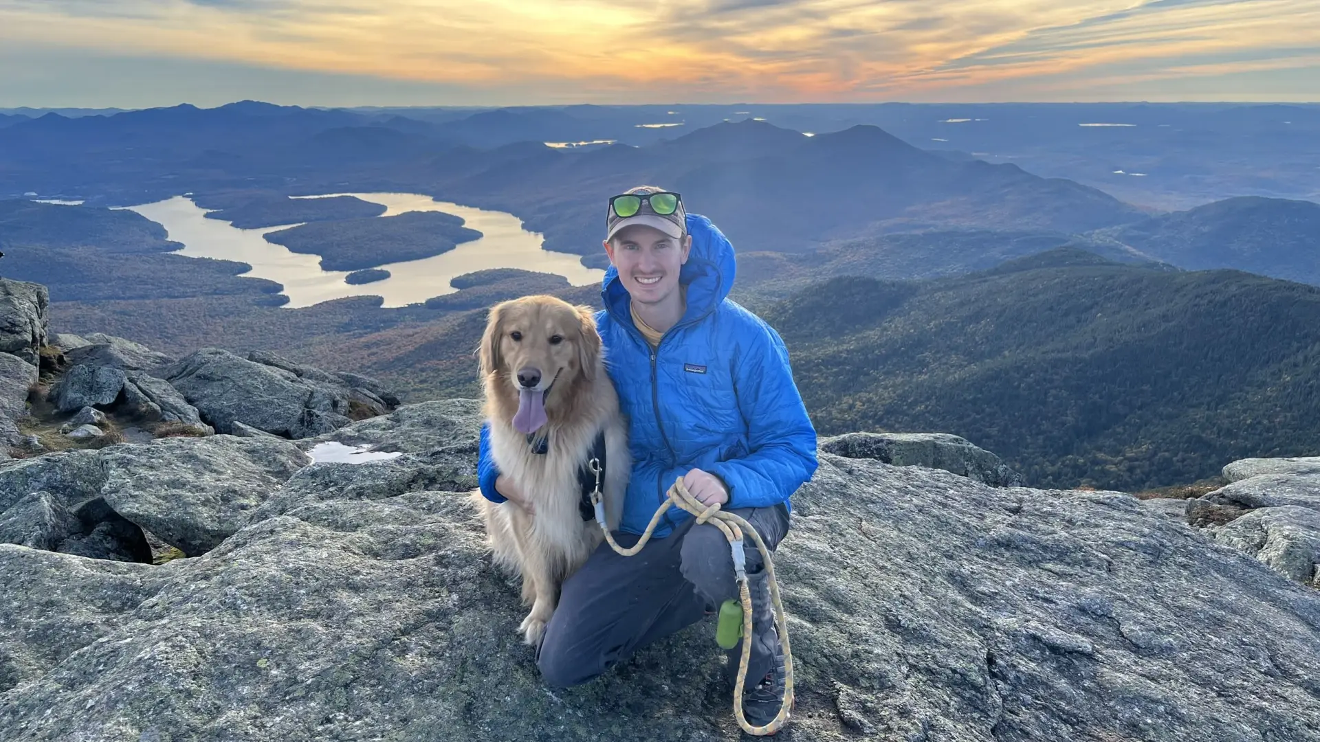 A man and his golden retriever sit atop a summit overlooking Lake Placid at sunset