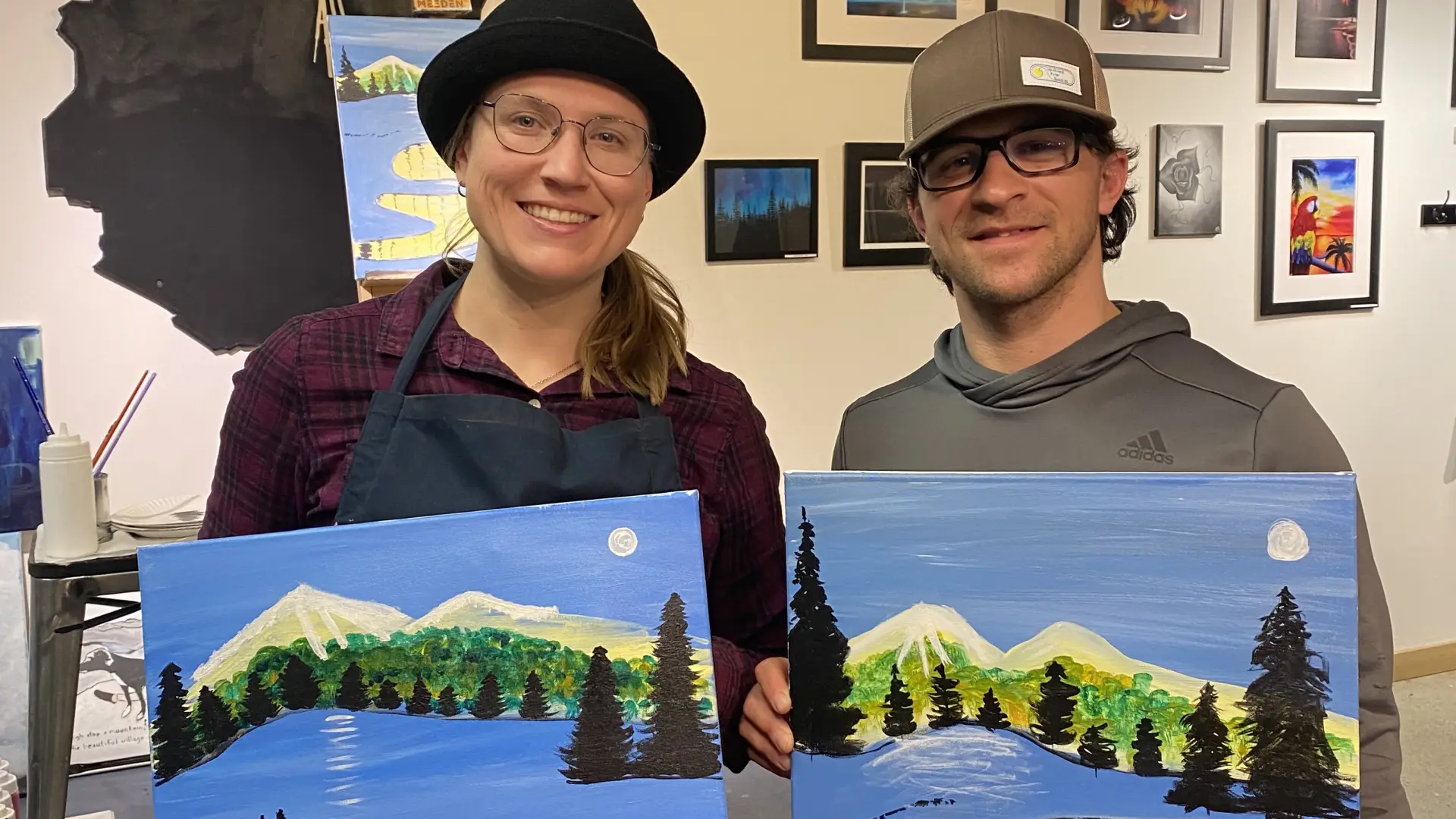 A man and a woman hold up paintings, each a version of the same image, from an art class.