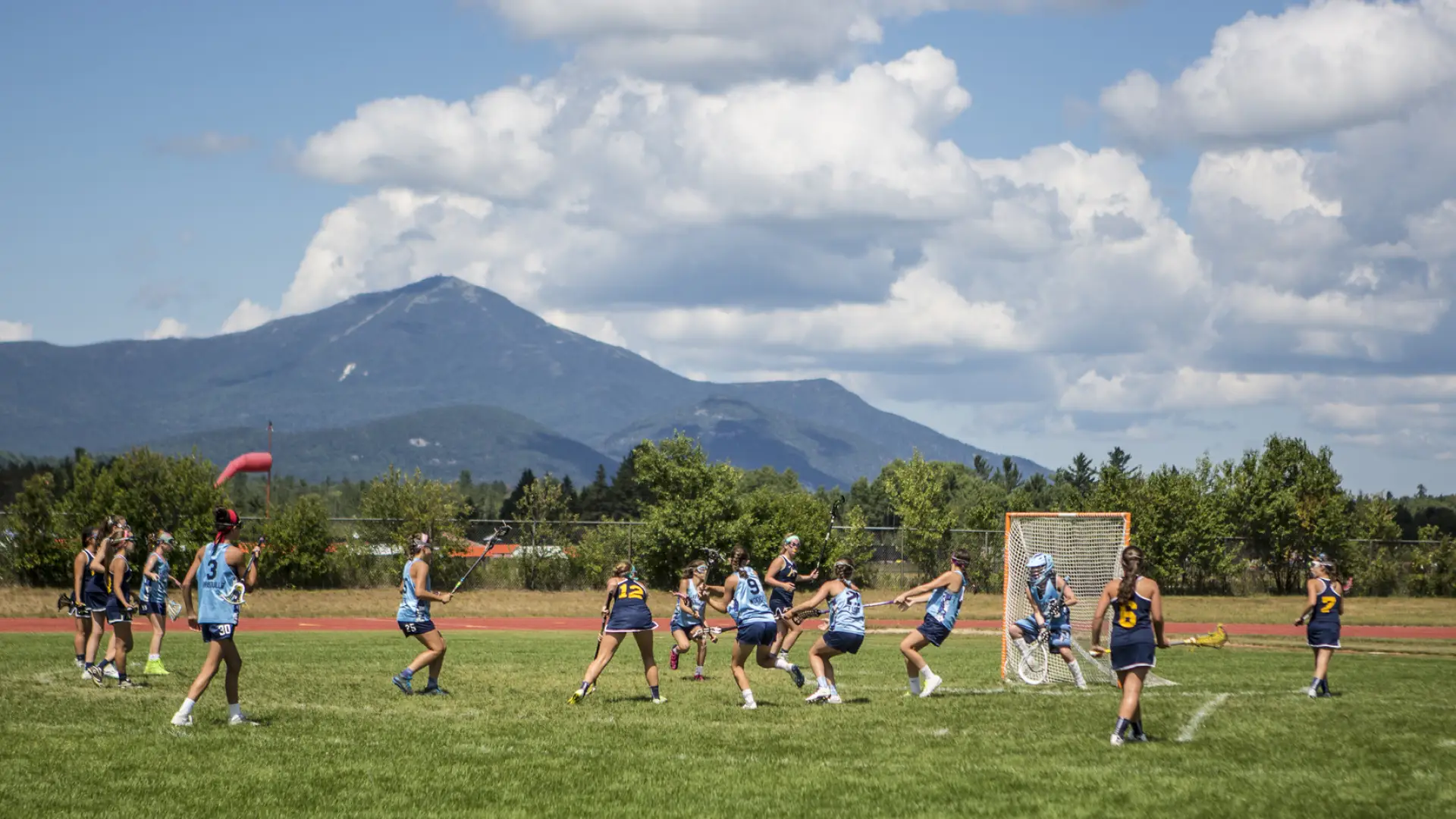 A group of athletes plays lacrosse on an outdoor field in Lake Placid, with high peaks scenery in the distance