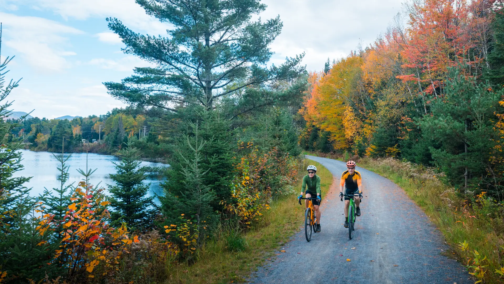 Two cyclists ride on a smooth gravel trail in fall, with a river to their right.