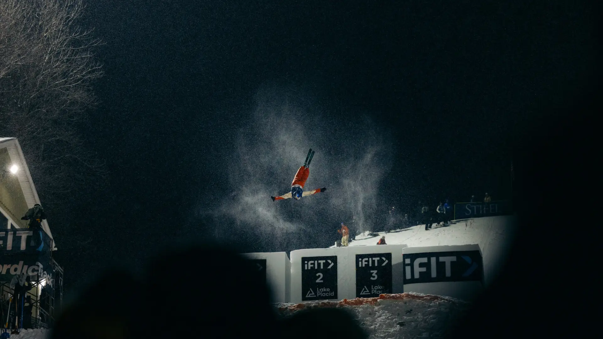 Freestyle aerialist skier performs a flip after skiing off a jump at a nighttime competition under bright lights while a crowd looks on