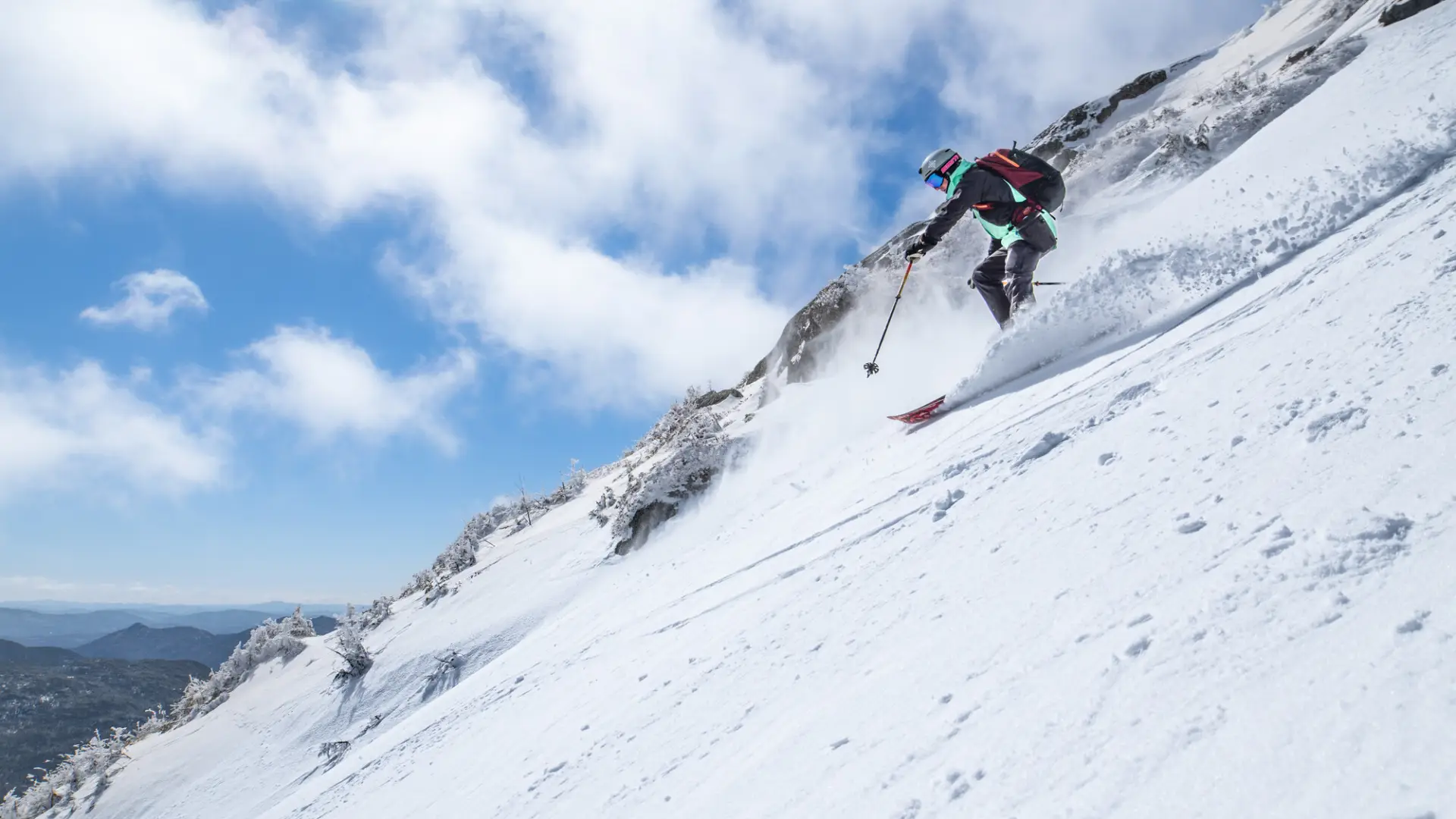 Skier in a red jacket descends a steep, snowy mountain slope under a bright blue sky. The scene conveys an exhilarating sense of adventure and speed.