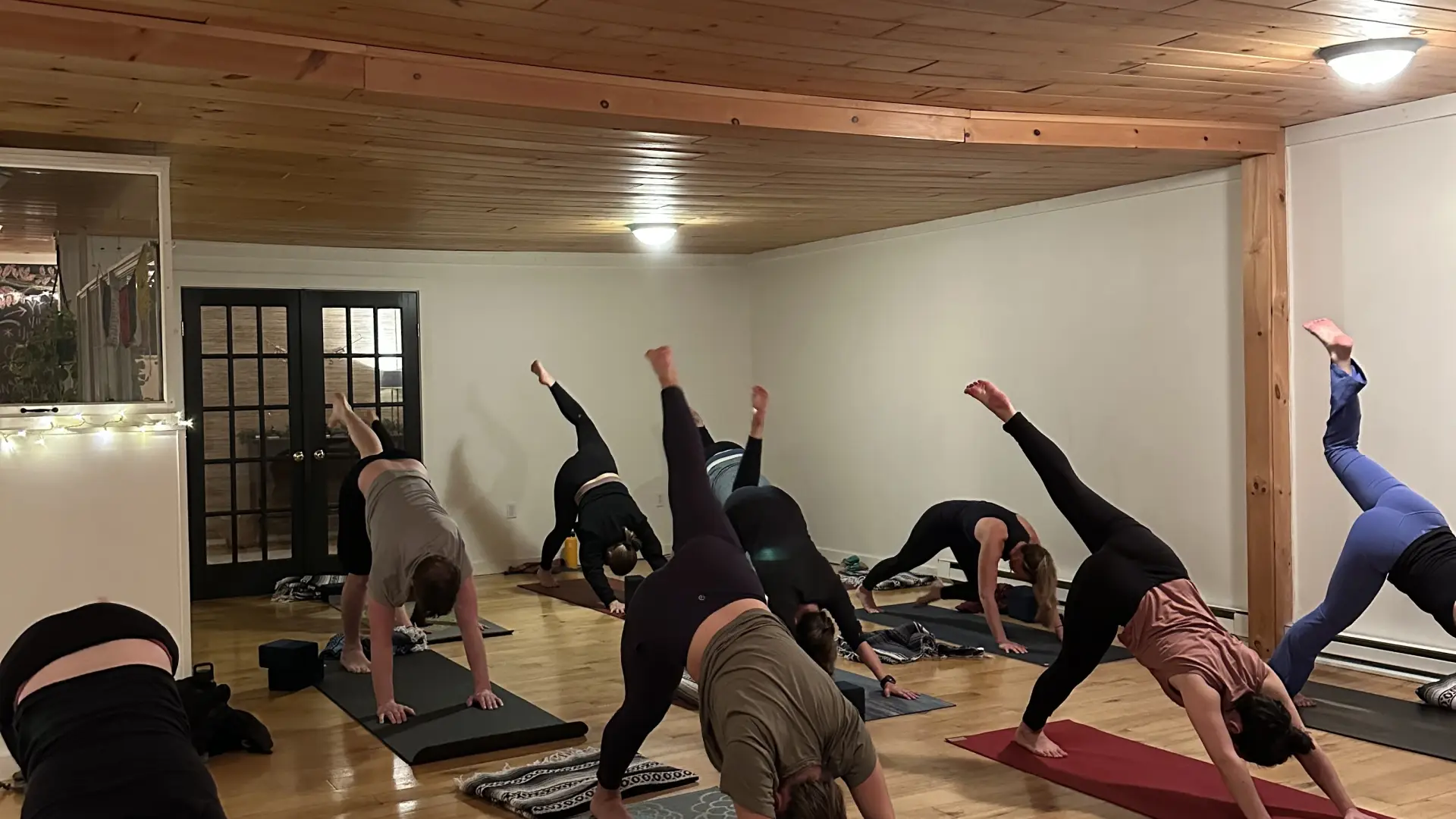 A yoga class in a studio with wood floors and a wood ceiling. Several people are doing a three-legged dog pose.