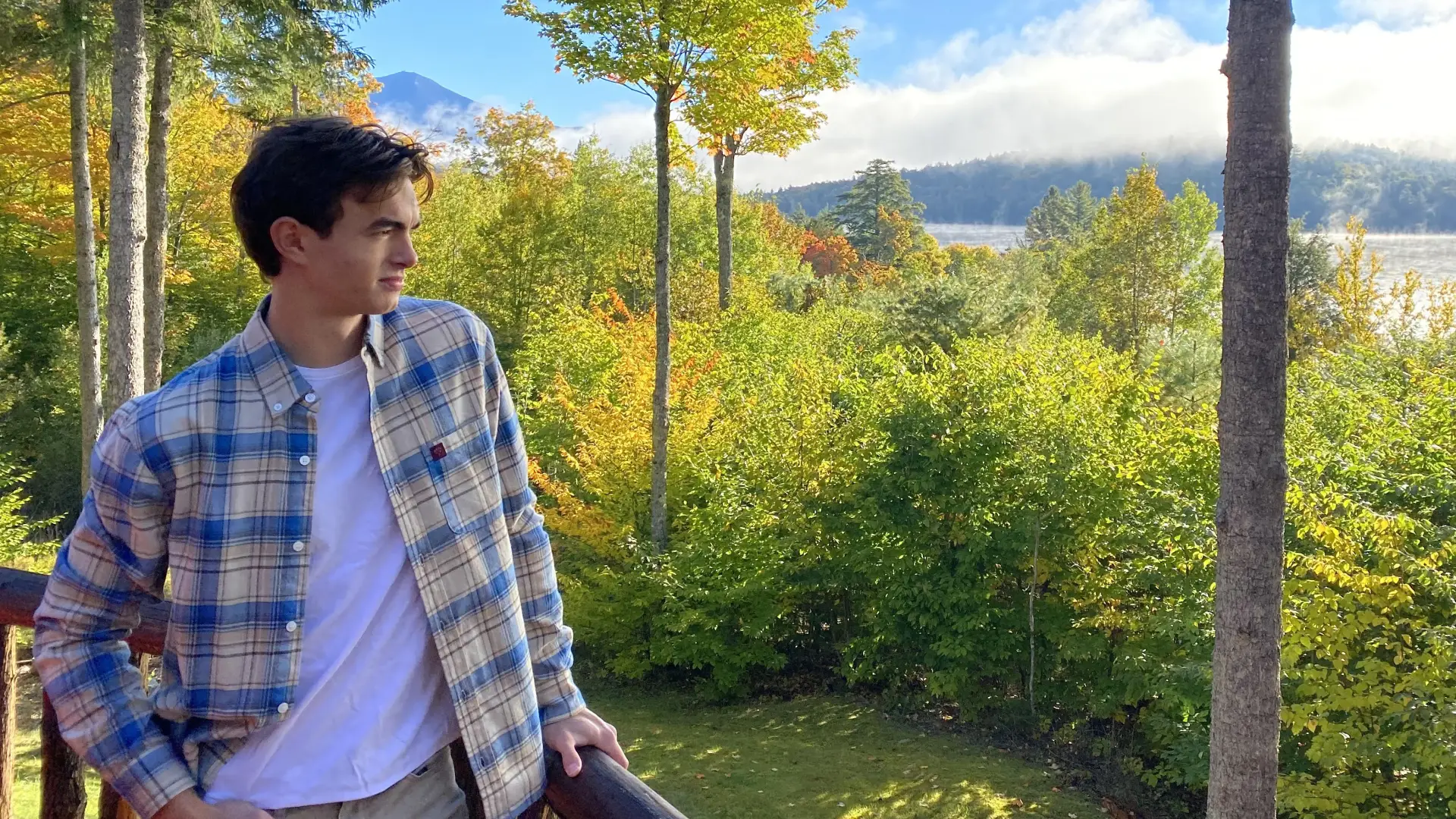 A young person wearing a flannel shirt poses and looks off into a forest glowing with golden light