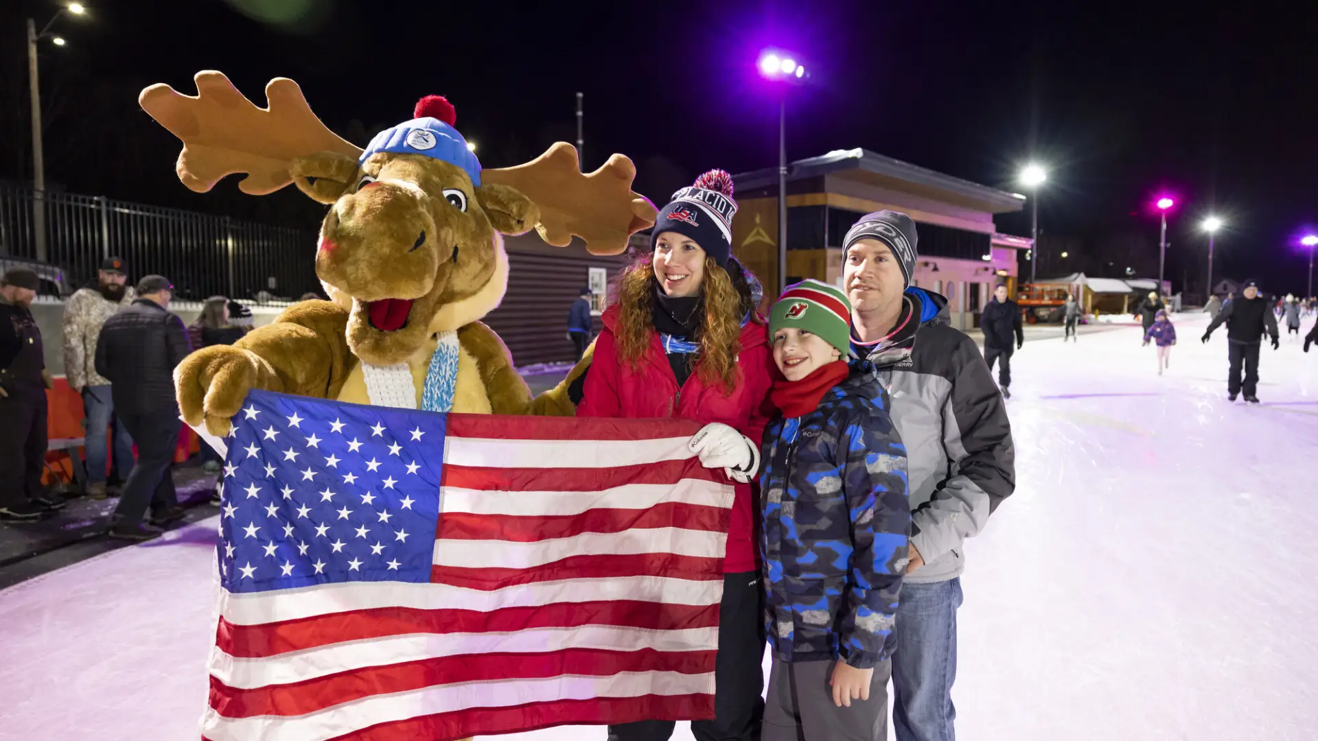 Family stands with moose mascot holding American flag on Olympic Speed Skating Oval at night taking group picture