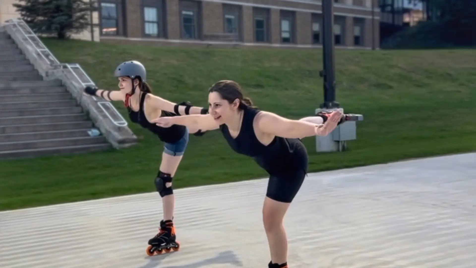 Two women inline skate side-by-side in figure skating poses, skating on one leg.