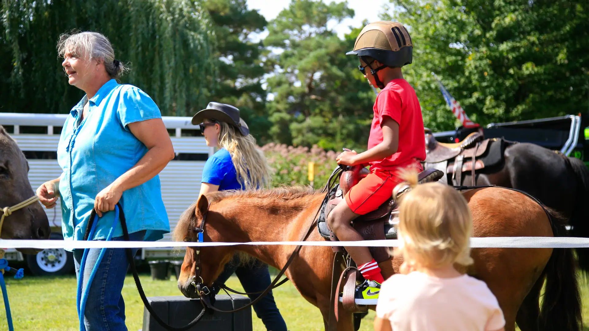 Young boy dressed all in red wearing a black helmet rides a small horse led by an older woman in a blue shirt while other children look on