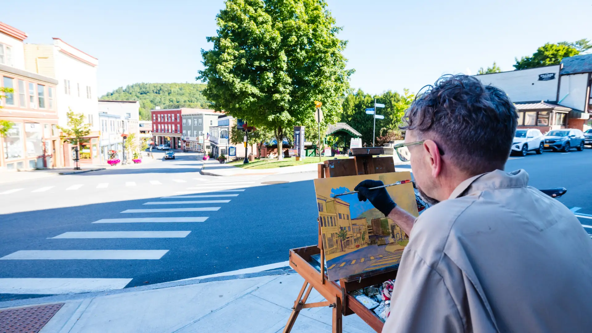 An artist stands at an easel on a sunny sidewalk, painting a plein air landscape of Saranac Lake.