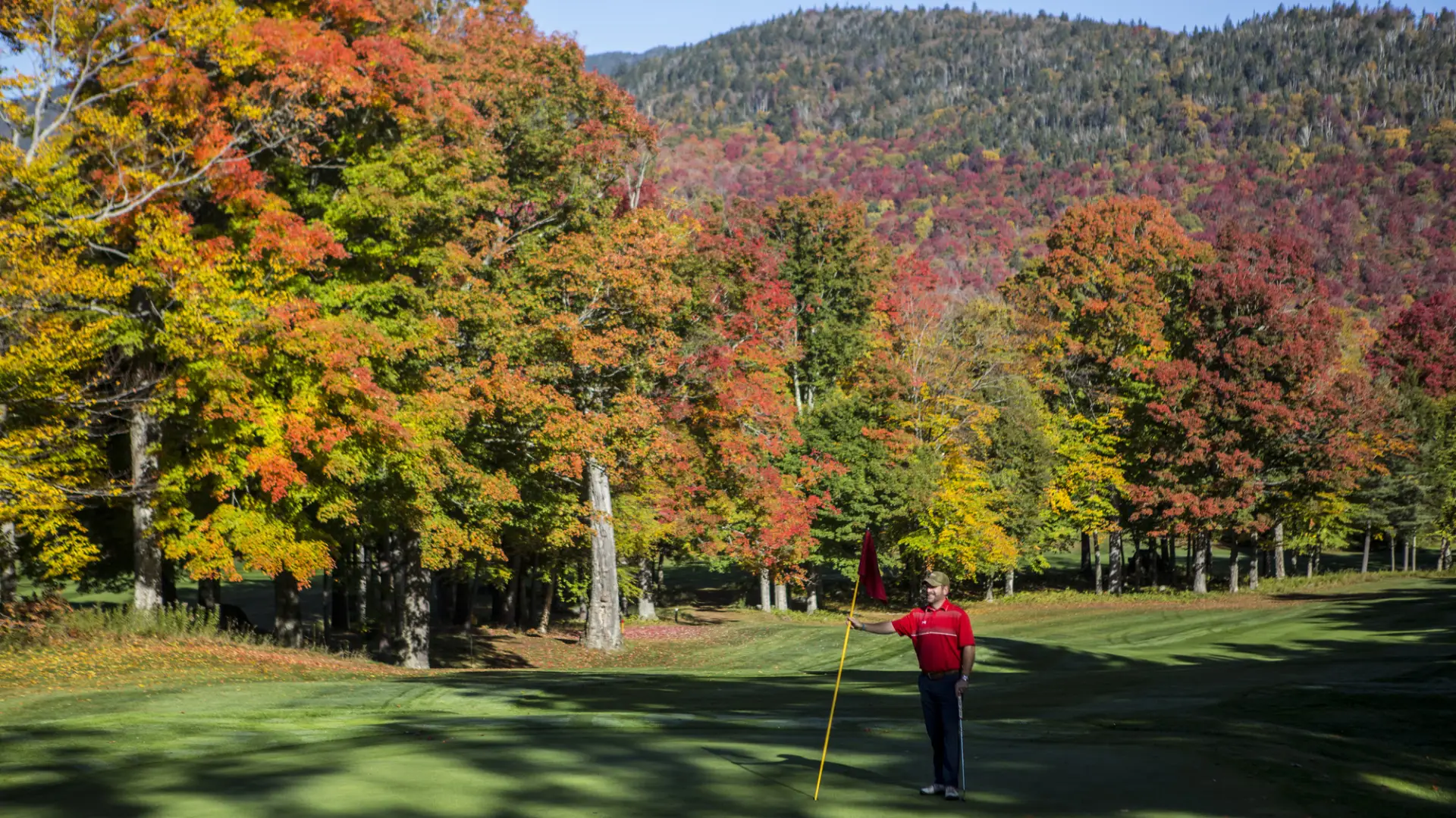 A man standing on the golf course holding a flag and smiling with beautiful fall leaves surrounding him.