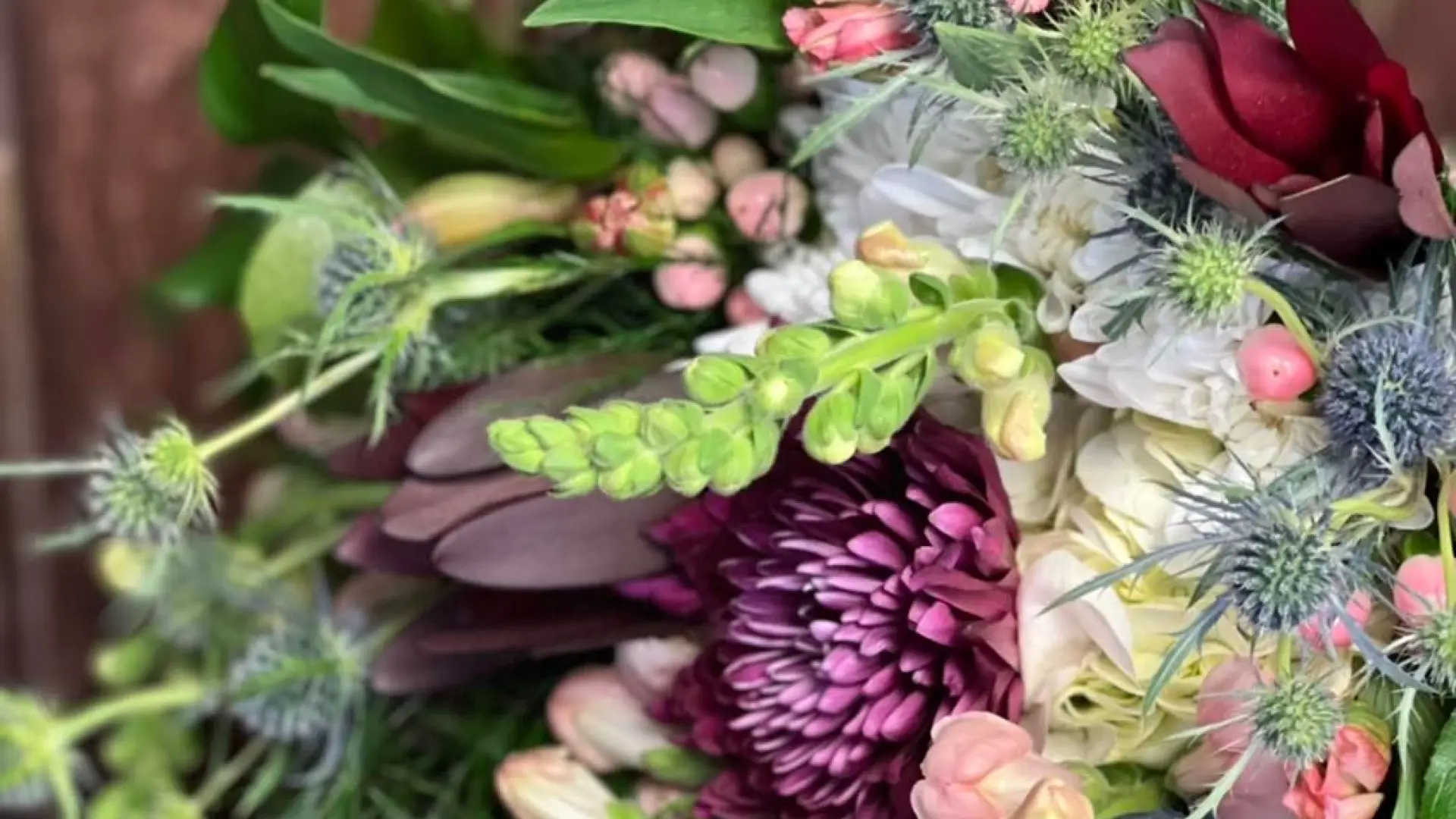 A bouquet of pink snap dragons, blue sea holly, and and burgundy daisies surrounded with assorted foliage accents lays on a table.
