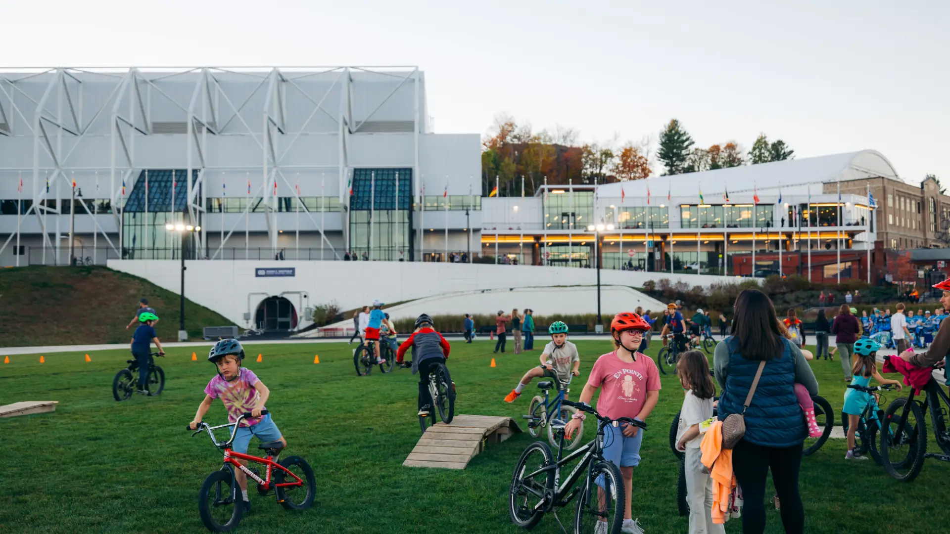 Kids ride on small mountain bike features on grassy area of Olympic Oval in Lake Placid with Olympic Center in background