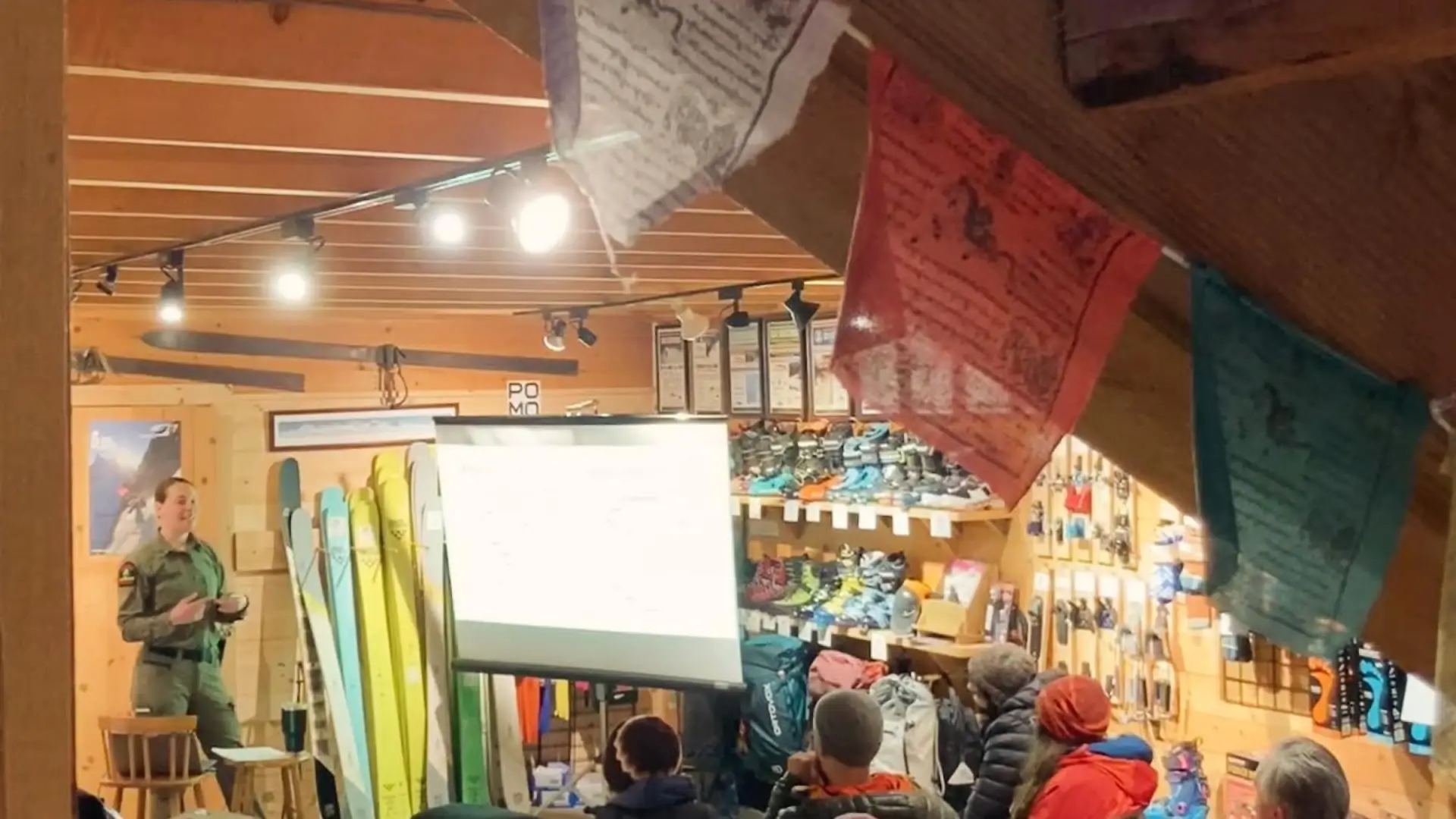 Forest Ranger gives a presentation next to a white screen to a room of sitting people with prayer flags hanging from the ceiling