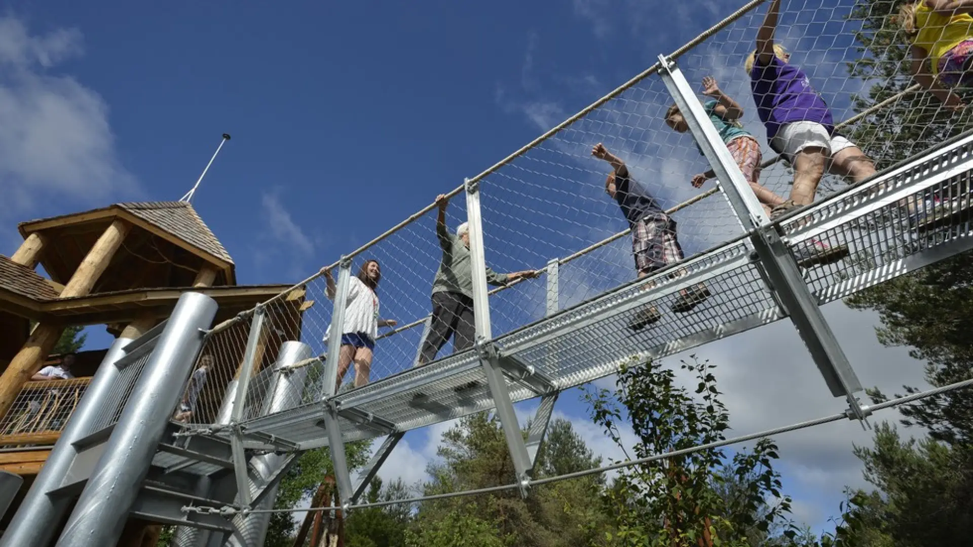 People crossing a suspended metal walkway between large wooden structures at The Wild Center in Tupper Lake NY 