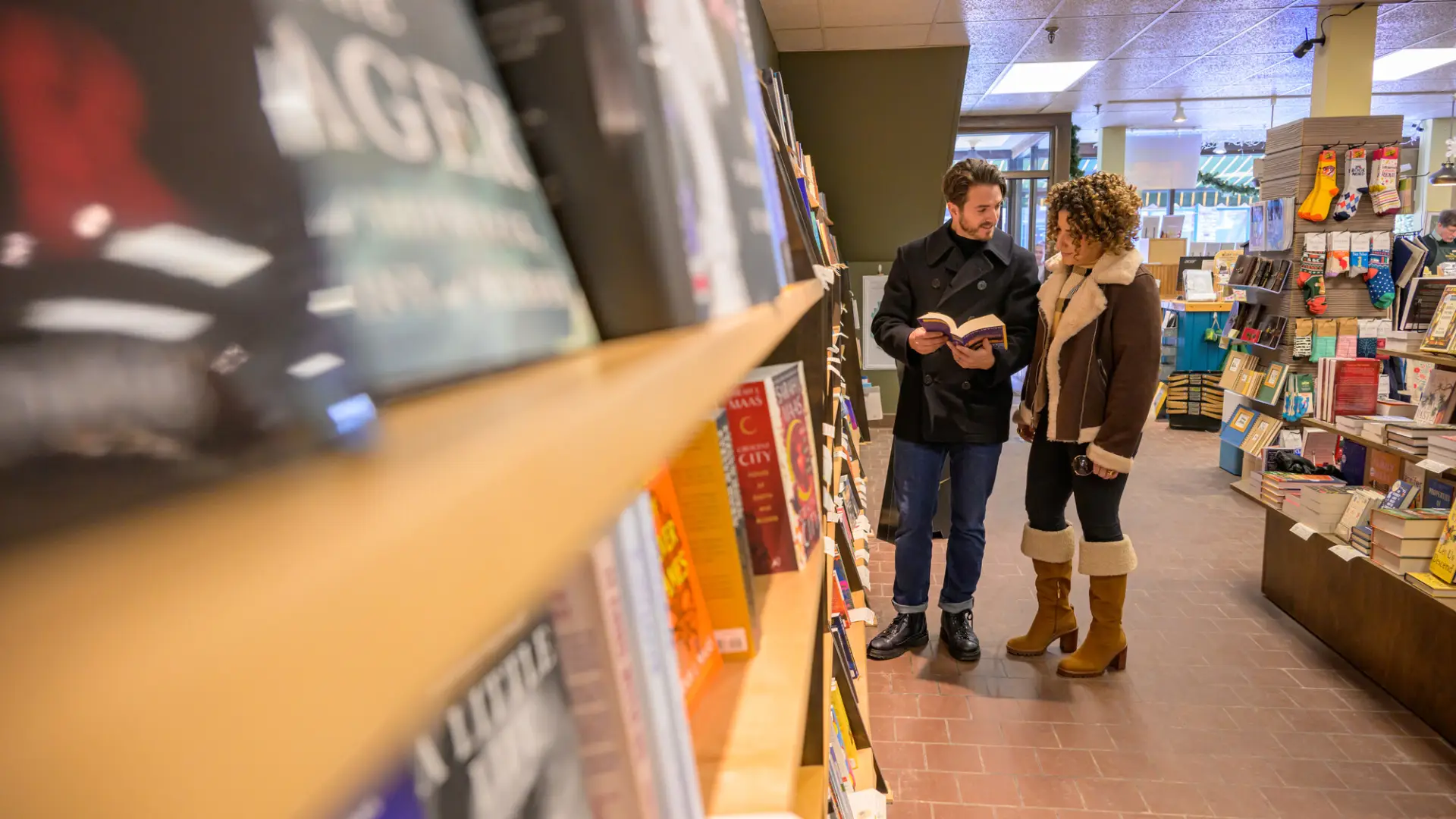 Couple in winter outerwear stands at bookshelves in bookstore looking at paperback
