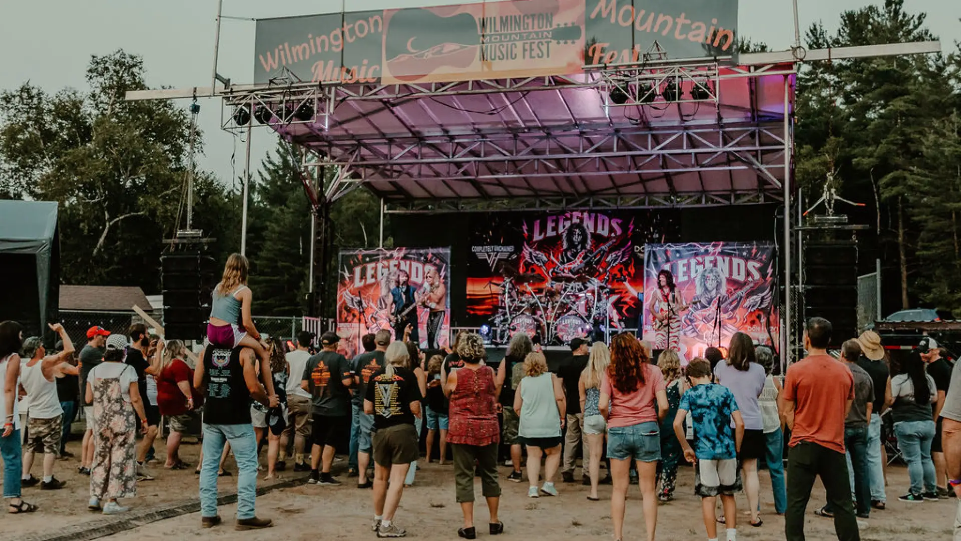 Crowd of people stand on dirt lawn in front of temporary music stage  in summer