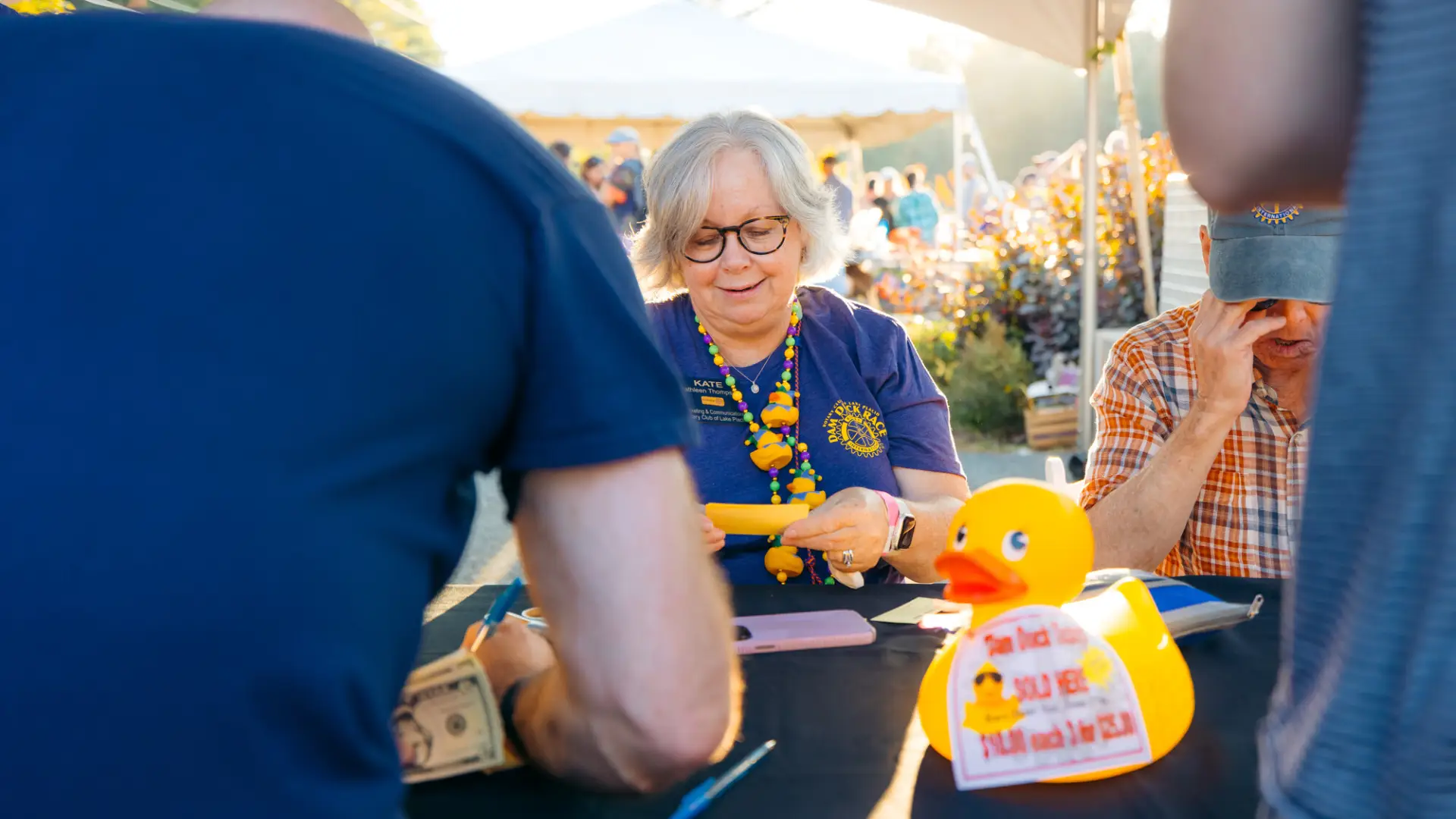 Woman with grey hair and glasses wearing blue Rotary shirt and duck accessories is seen selling something 