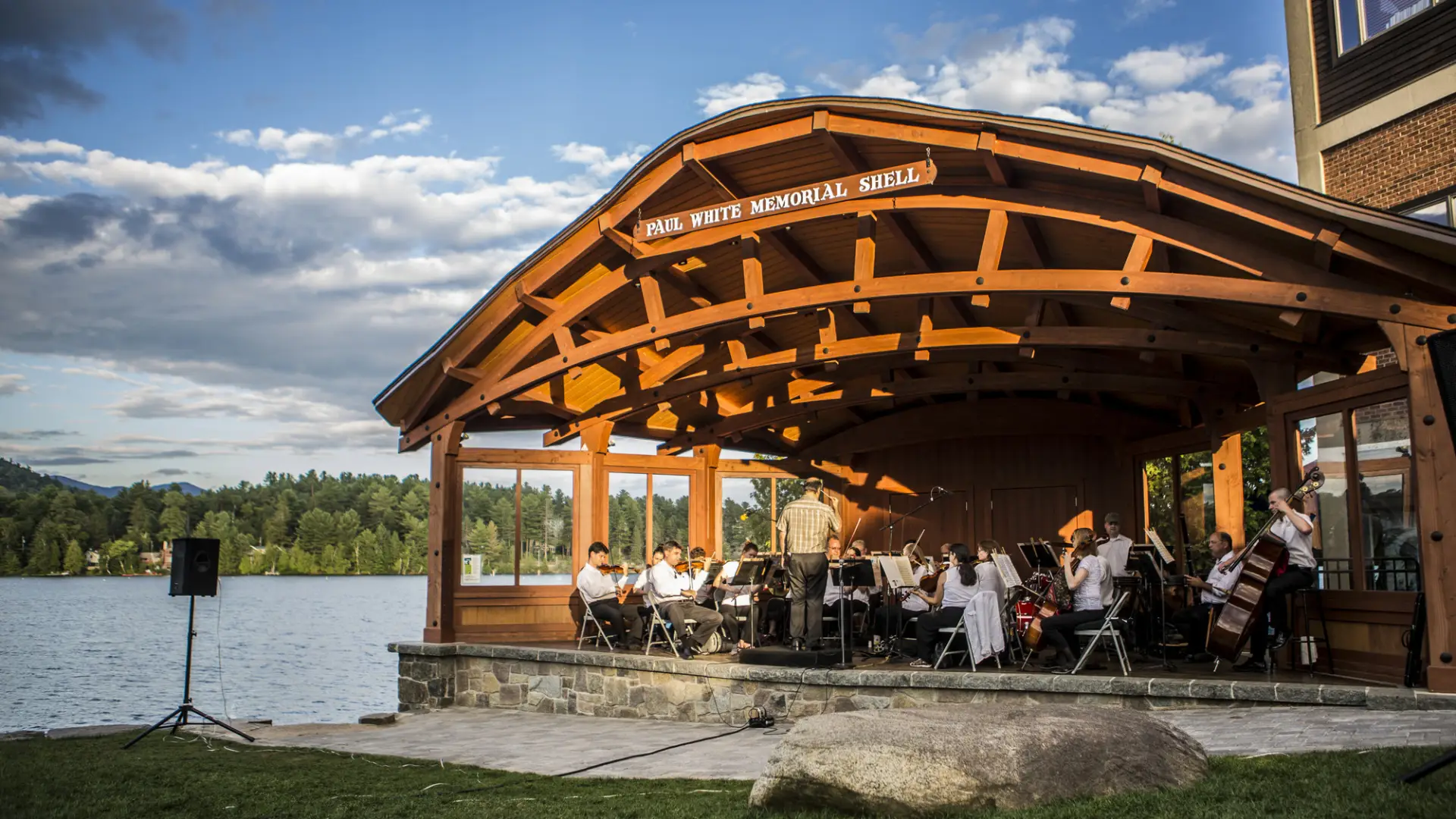 A sinfonietta playing instruments on a bandshell stage with a lake in the background.