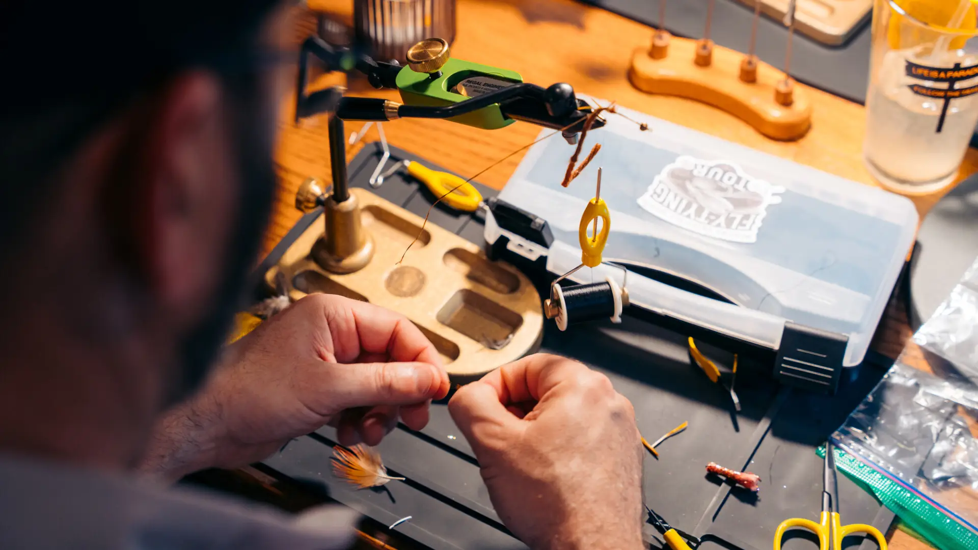 A person’s hands are shown as they meticulously tie a fly on a green and gold rotary vise at a wooden workbench.
