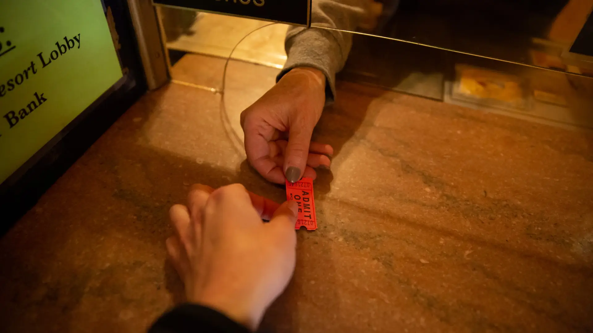 A close-up shot captures one hand passing a small "ADMIT ONE" ticket to another person across a counter at a box office.