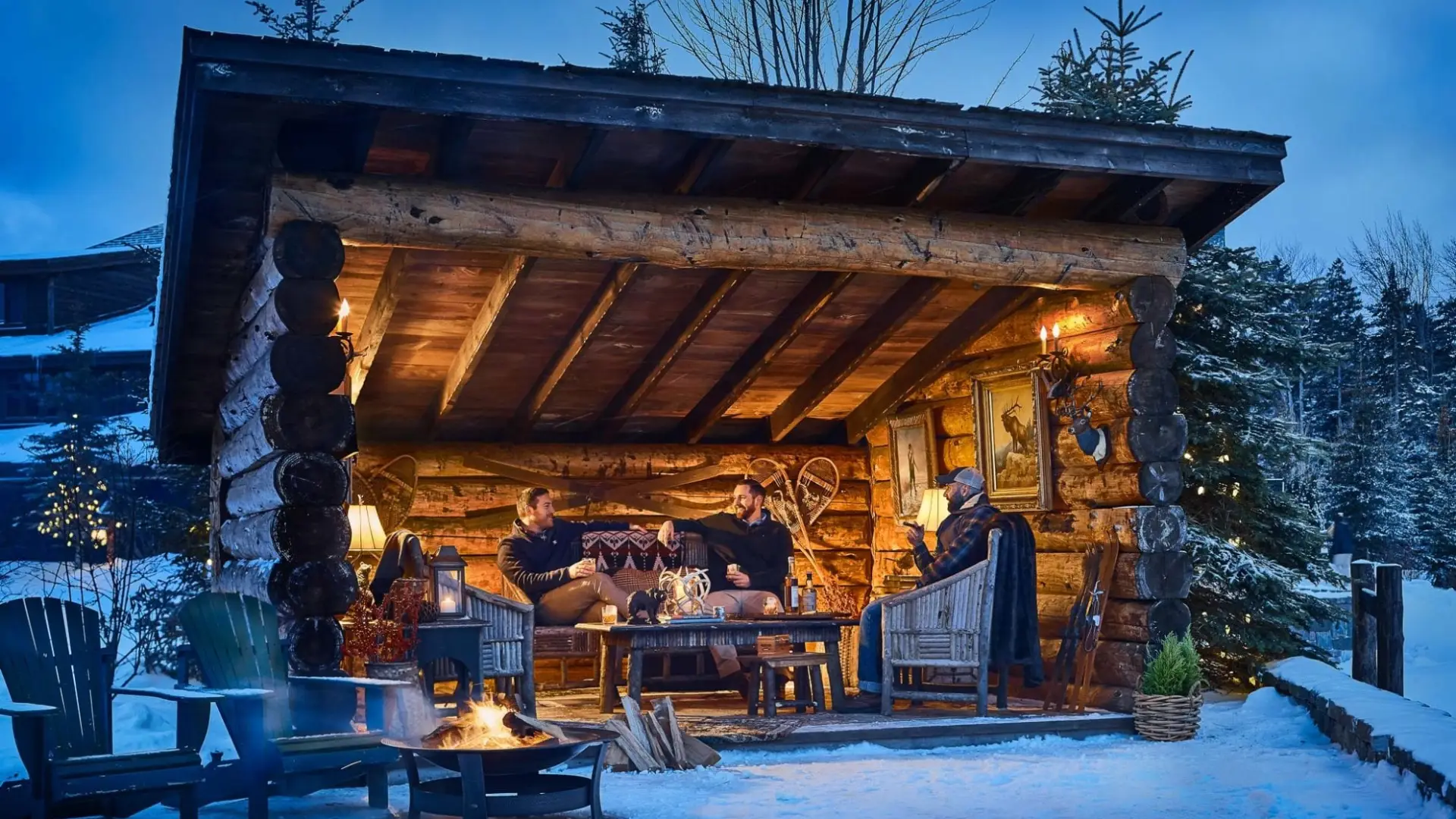 Two adults sit in a luxuriously appointed rustic lean-to on a snowy winter evening.