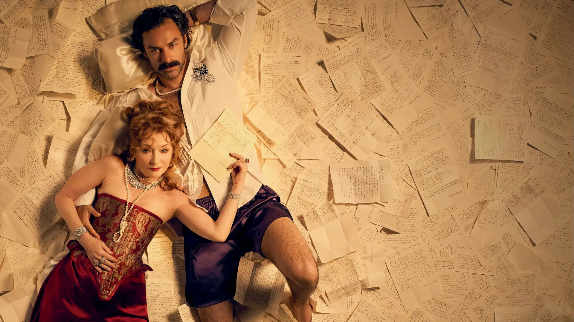 An actor in white shirt and shorts and actress in red corset dress lay together amidst a pile of papers