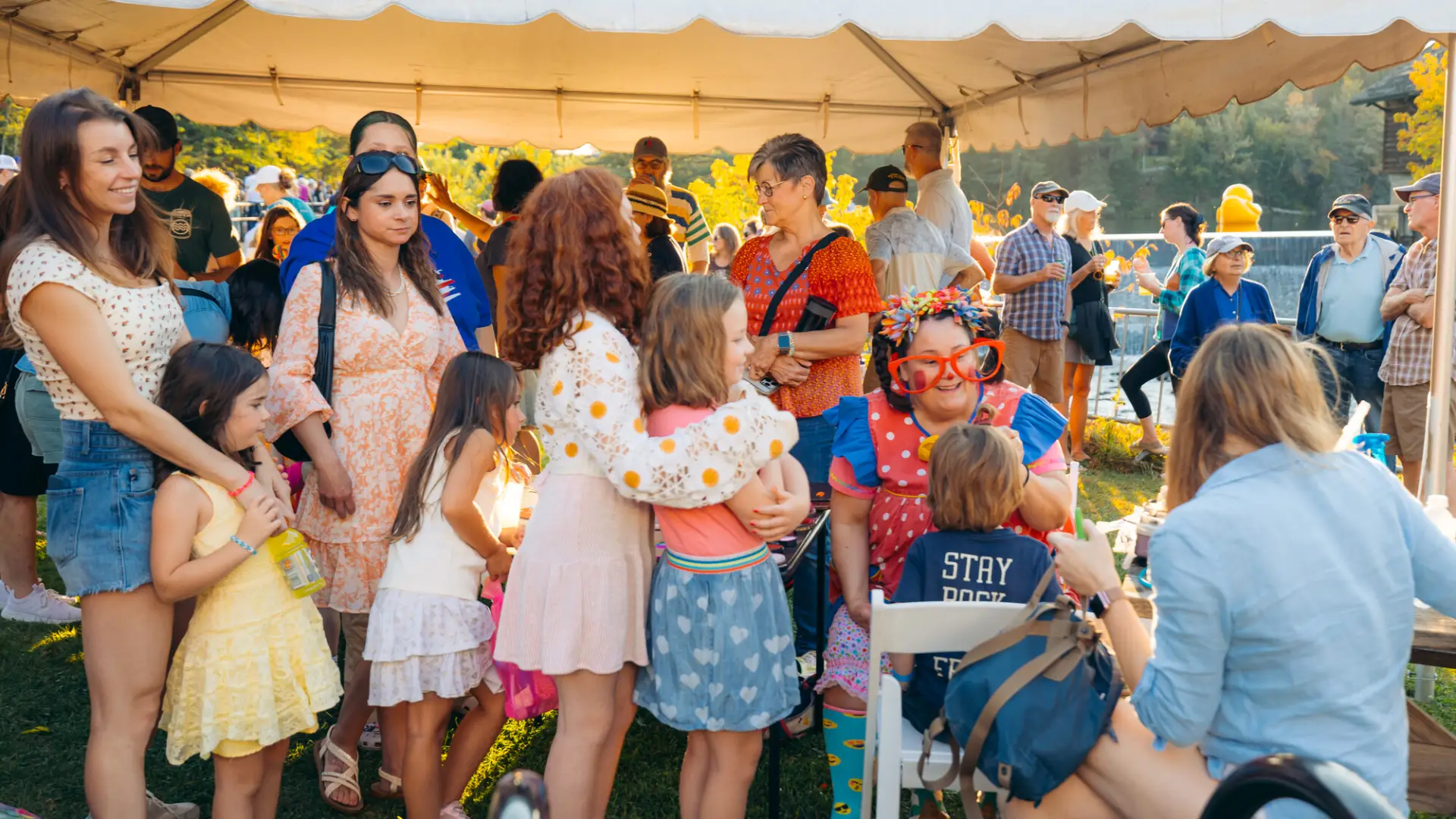 Children and parents wait in line looking on as seated clown paints the face of a child 