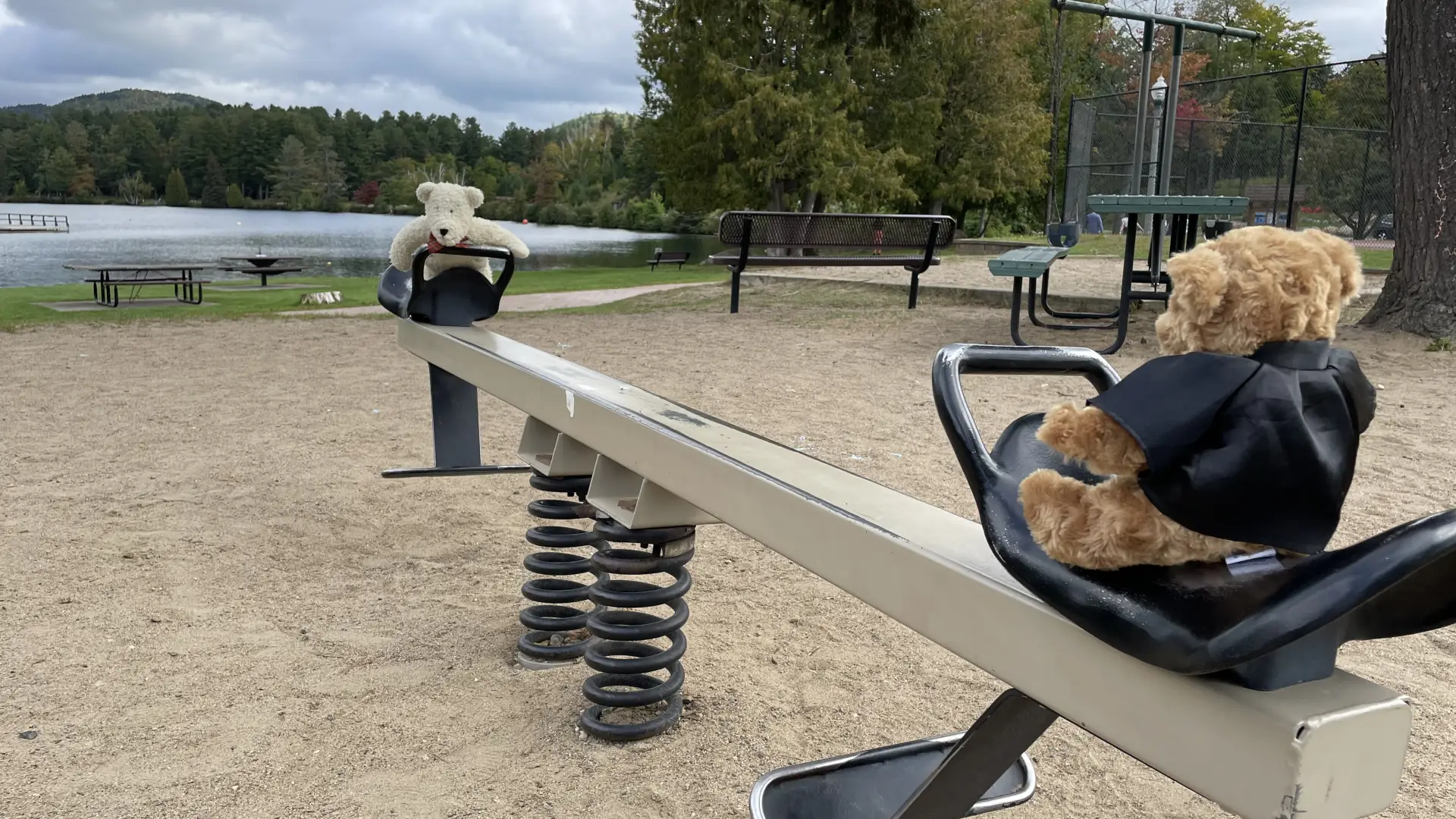 Two teddy bears sit on a teeter totter on a playground with a lake in the background.