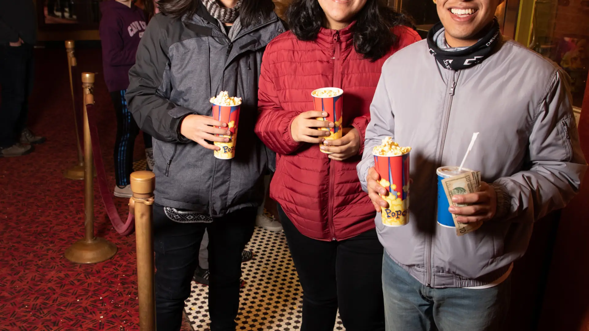 Three people stand in a movie theater lobby, smiling and holding buckets of popcorn.