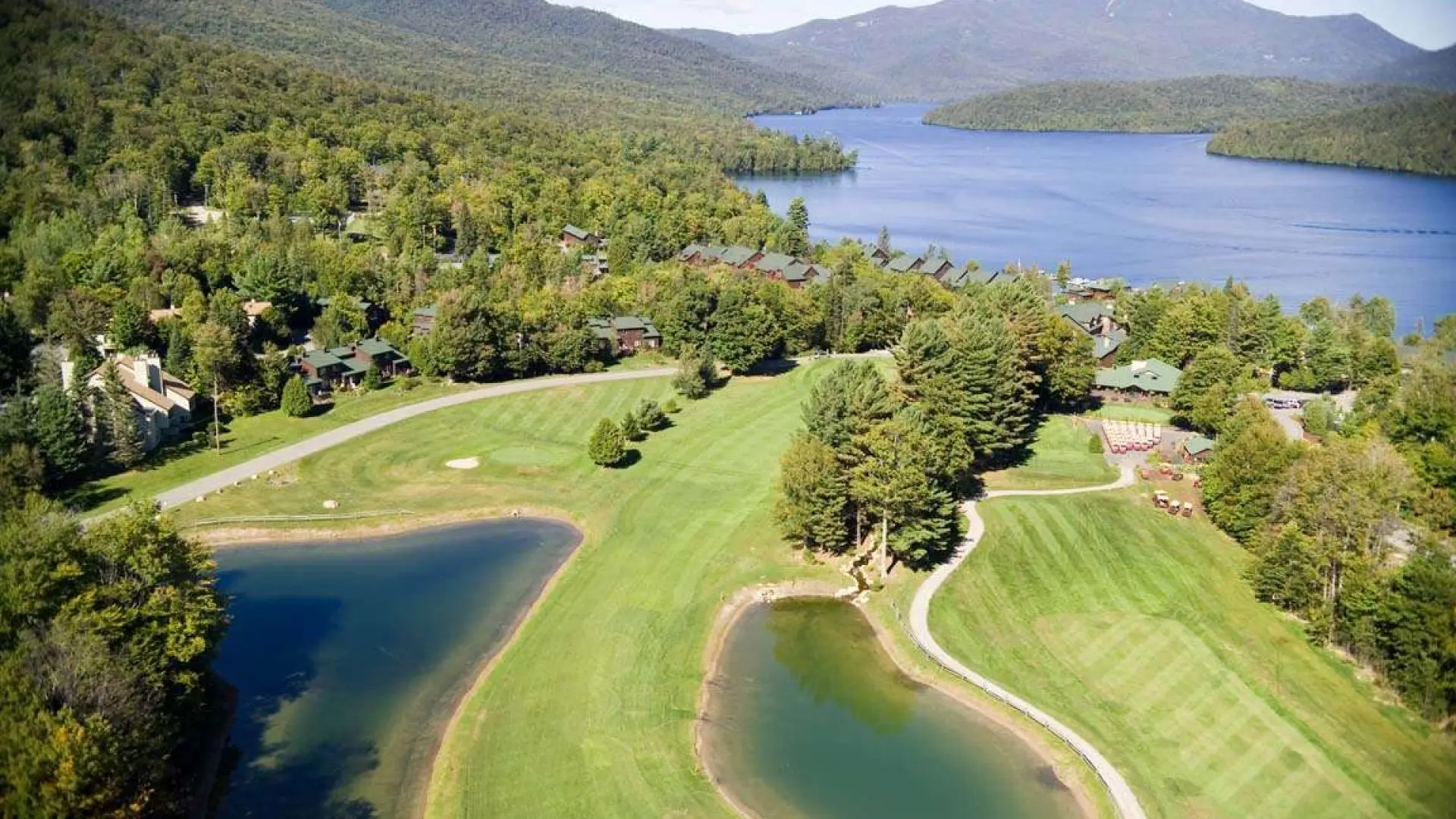An aerial view of the pristine grass of the course next to the deep blue of Mirror Lake and the mountains of the High Peaks surrounding.