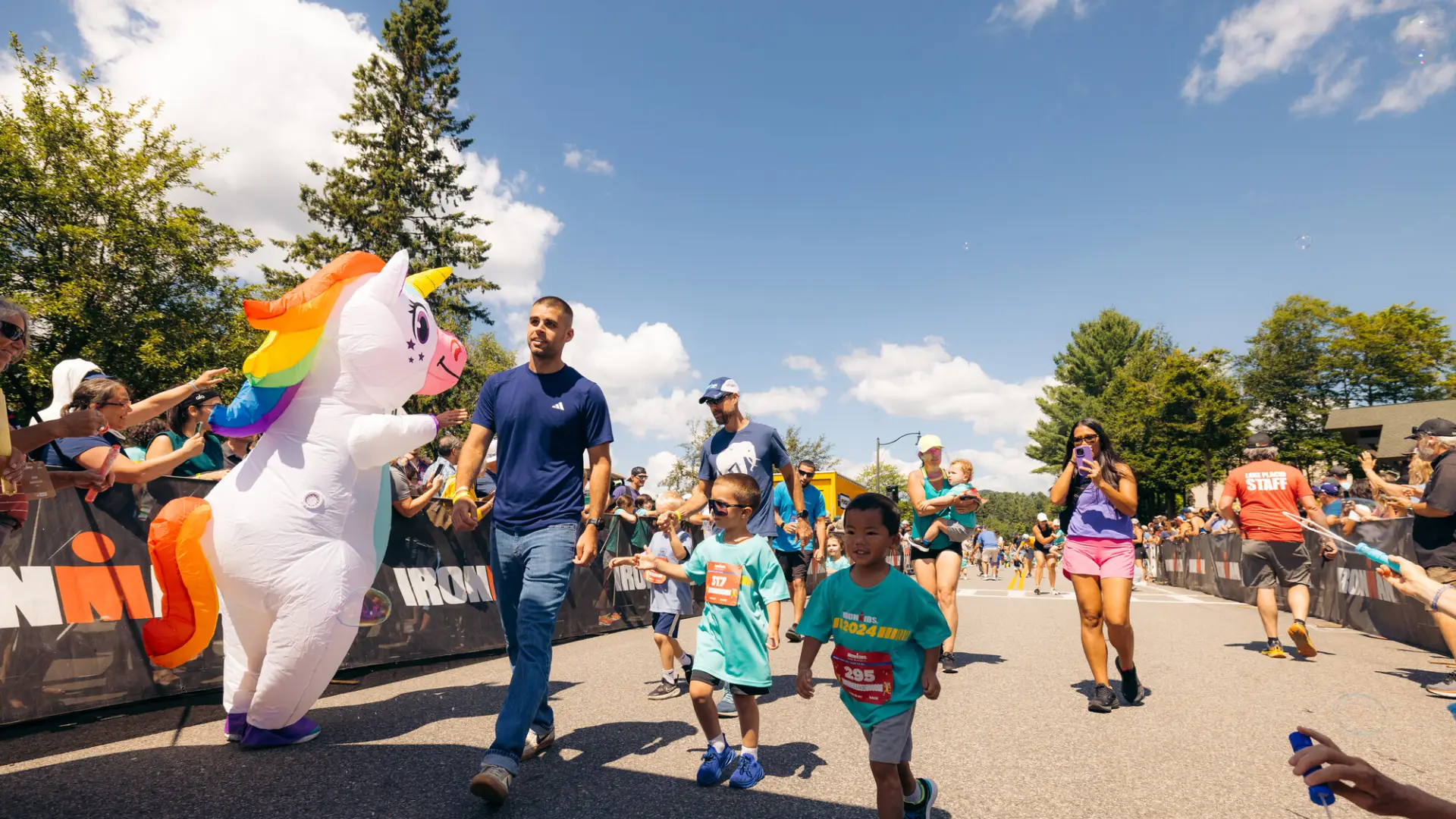 Group of kids and parents, some filming, walk in Ironkids race course past inflatable unicorn mascot