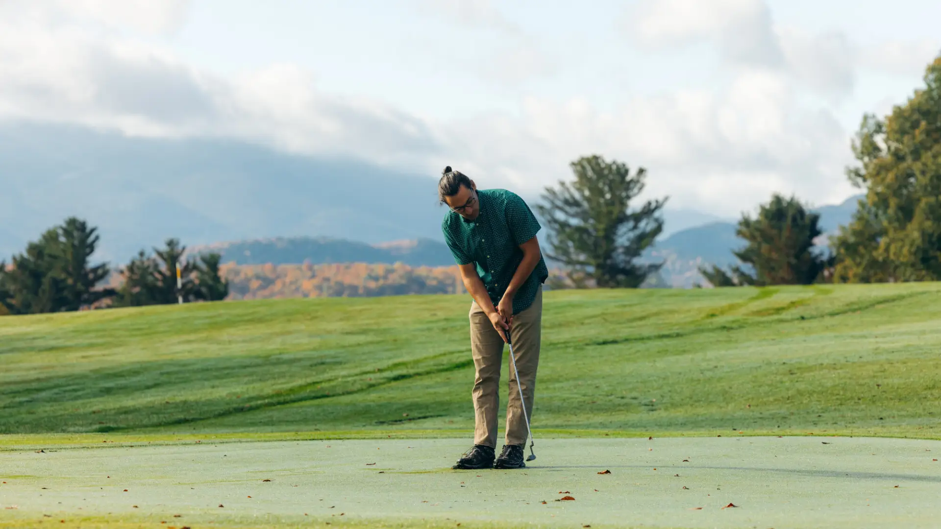 A golfer putting a ball on the green with mountain vistas in the background.