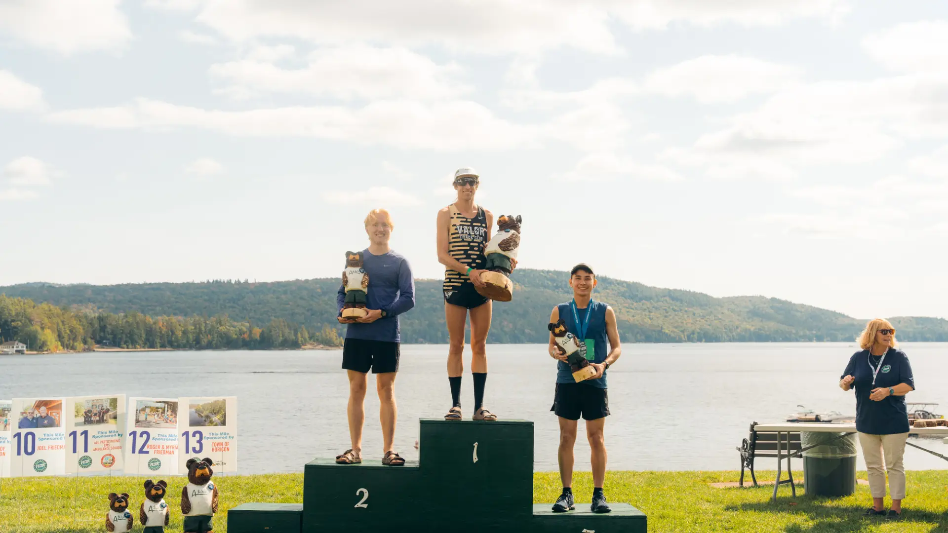 Three male runners stand on a three-tiered winner's podium by a lake, each holding a carved bear trophy.
