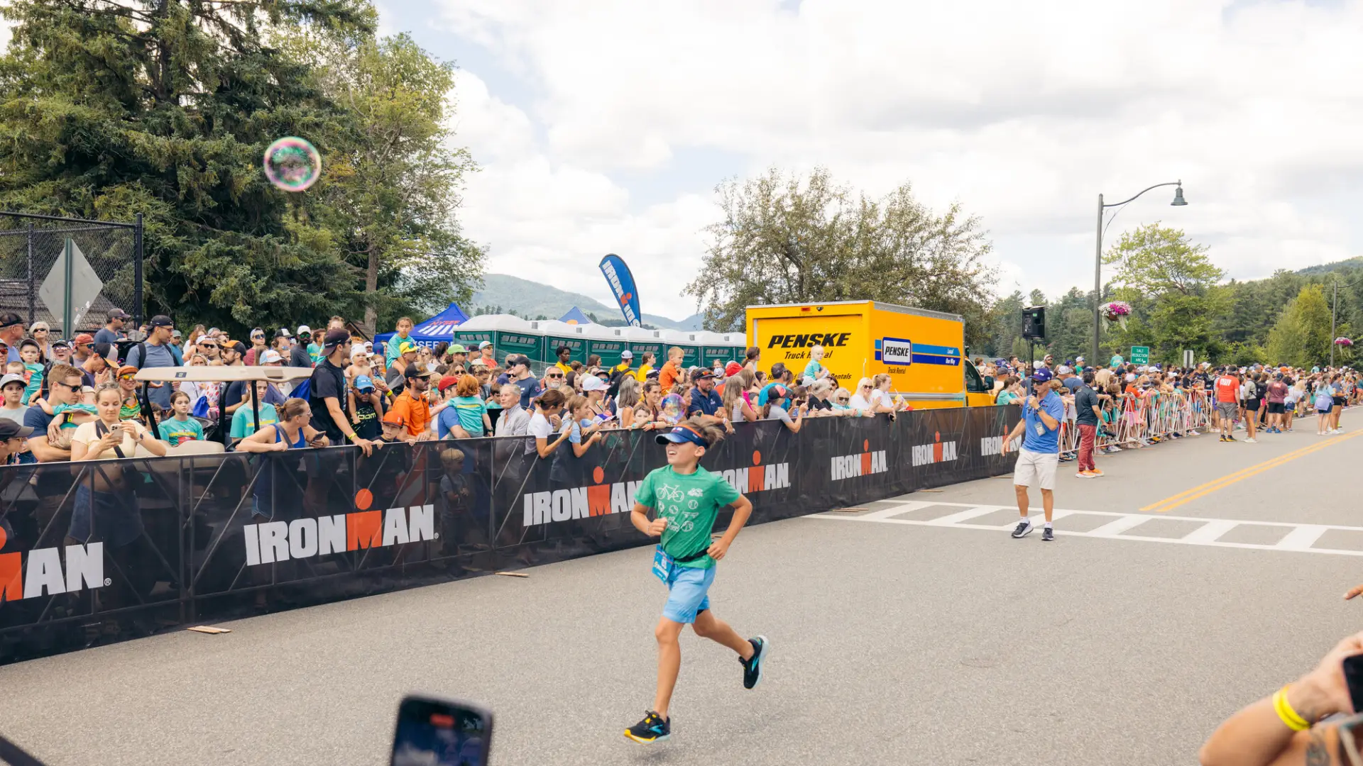 Child in teal shirt, blue shorts, and visor runs on Ironkids course while spectators cheer from behind Ironman fencing