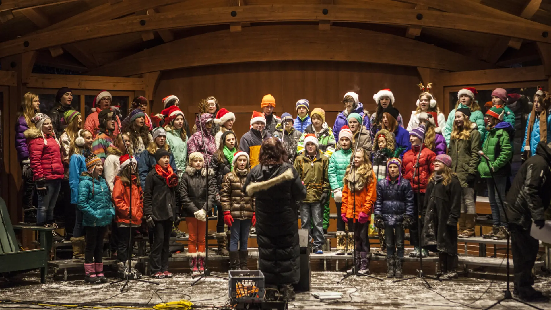 Several rows of children in winter and Santa hats sing in a lit, outdoor bandshell.