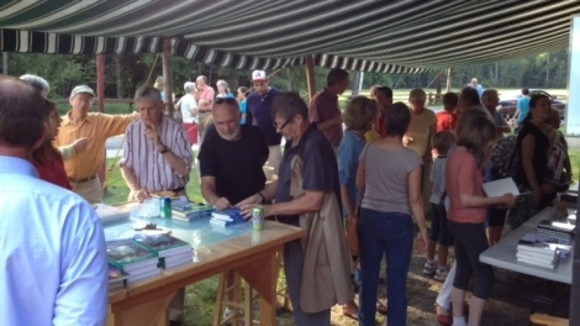 A group of people gather under a striped awning, browsing books displayed on a table.