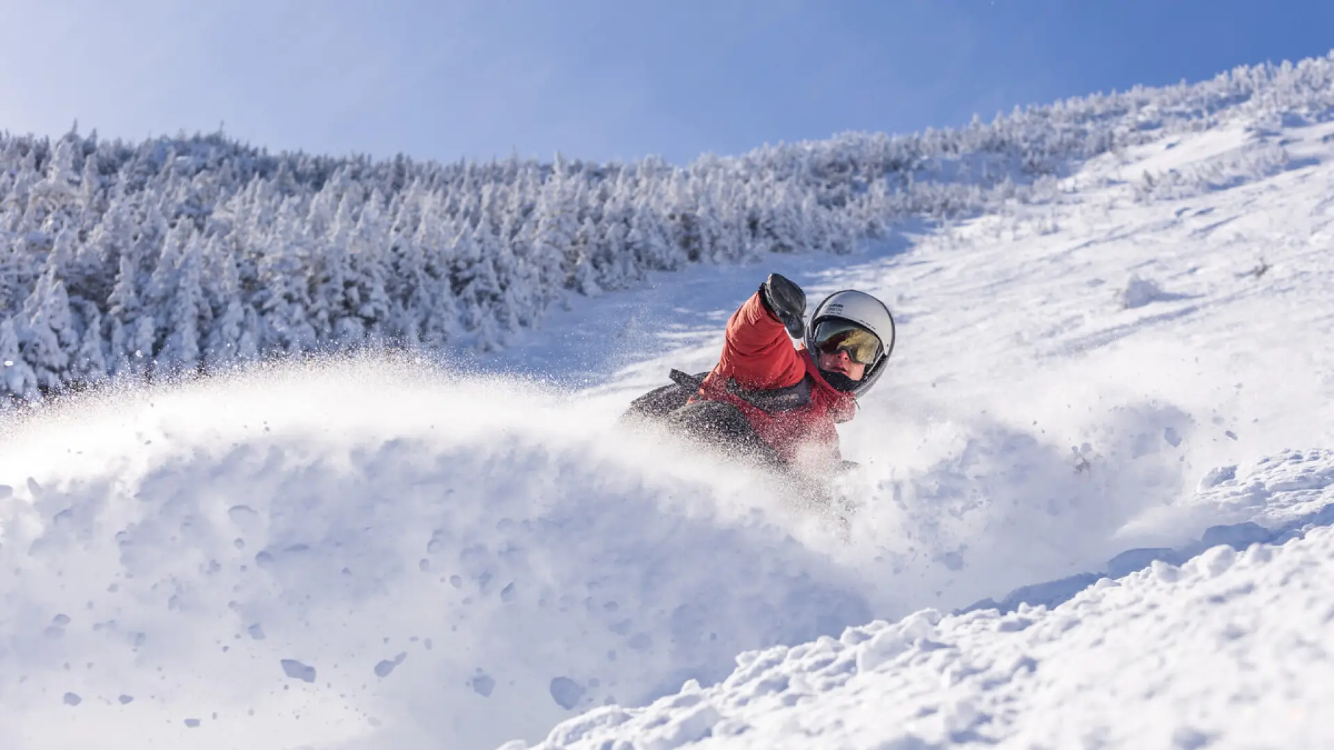Snowboarding in red jacket, helmet, and goggles sprays snow as they descend a steep, powdery, ungroomed slope