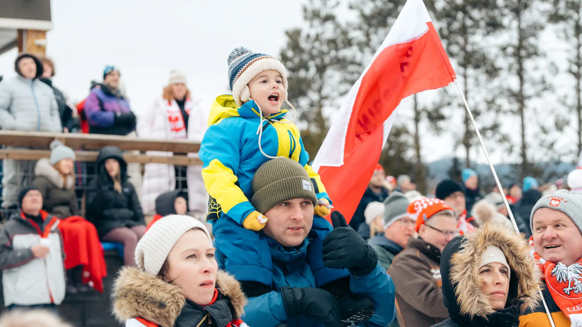 A small child sits on the shoulders of an adult male, in a crowd cheering at a winter outdoor sports event.