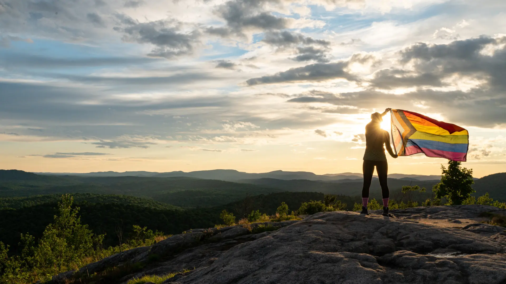 A person stands on a rocky hilltop, holding a brightly colored pride flag against a scenic sunset. The sky is dramatic with scattered clouds.