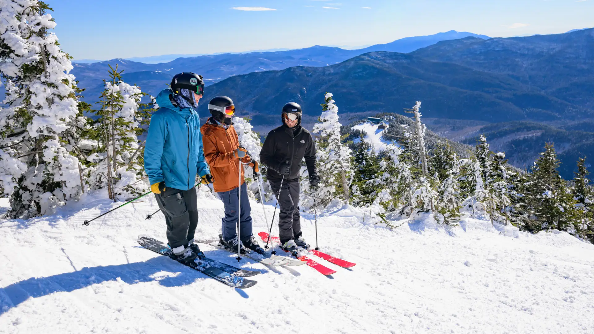 Three skiers stand at side of ski run in ski gear talking with snow encrusted trees beside them and snowy mountains in the distant background